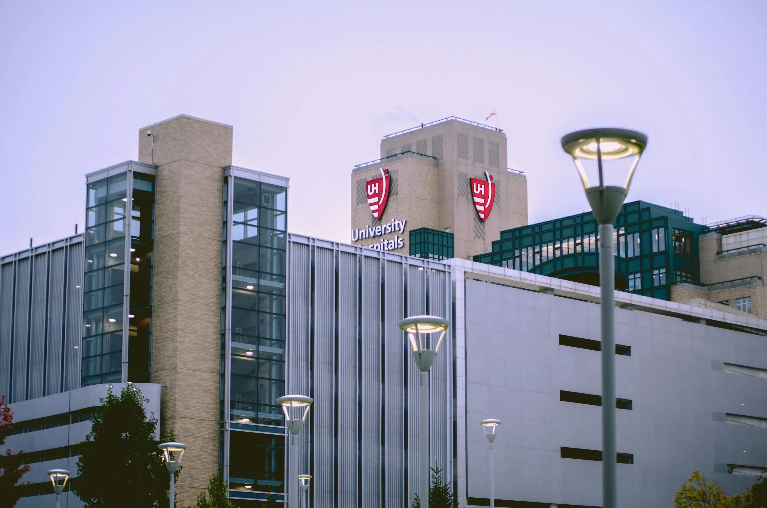 A modern hospital building with a sign that reads 'University Hospitals' and the UH logo on the top of the building, with streetlights and trees in the foreground.