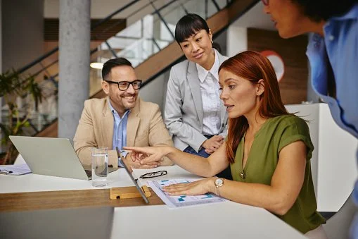 A meeting room with a diverse group of professionals, mostly women, sitting around a conference table, some taking notes and others listening attentively.