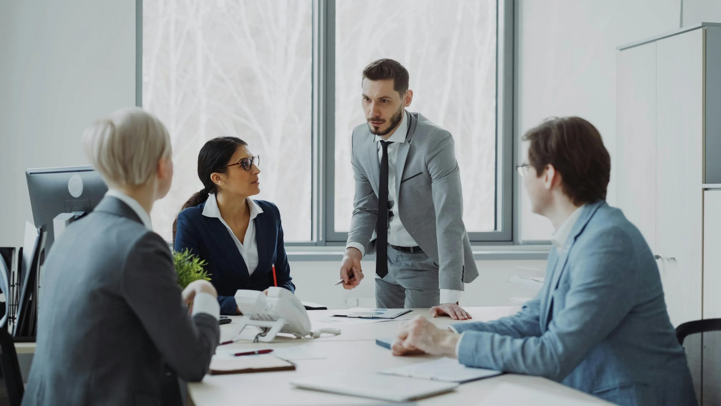 Four business professionals engaged in a meeting in an office conference room. A man in a grey suit is standing, leaning forward on the table, speaking to three seated colleagues, two women and one man, all in professional attire.