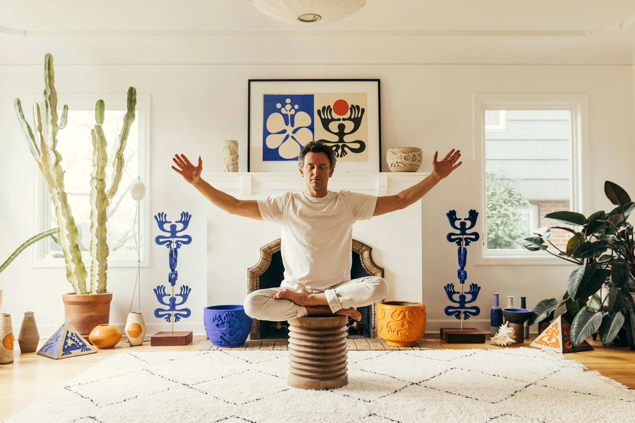 A man sitting cross-legged on a wooden platform in a beautifully decorated living room, practicing meditation with arms outstretched. The room features a white wall with framed artwork, various colorful pottery and sculptures, plants, and natural light coming through the windows.