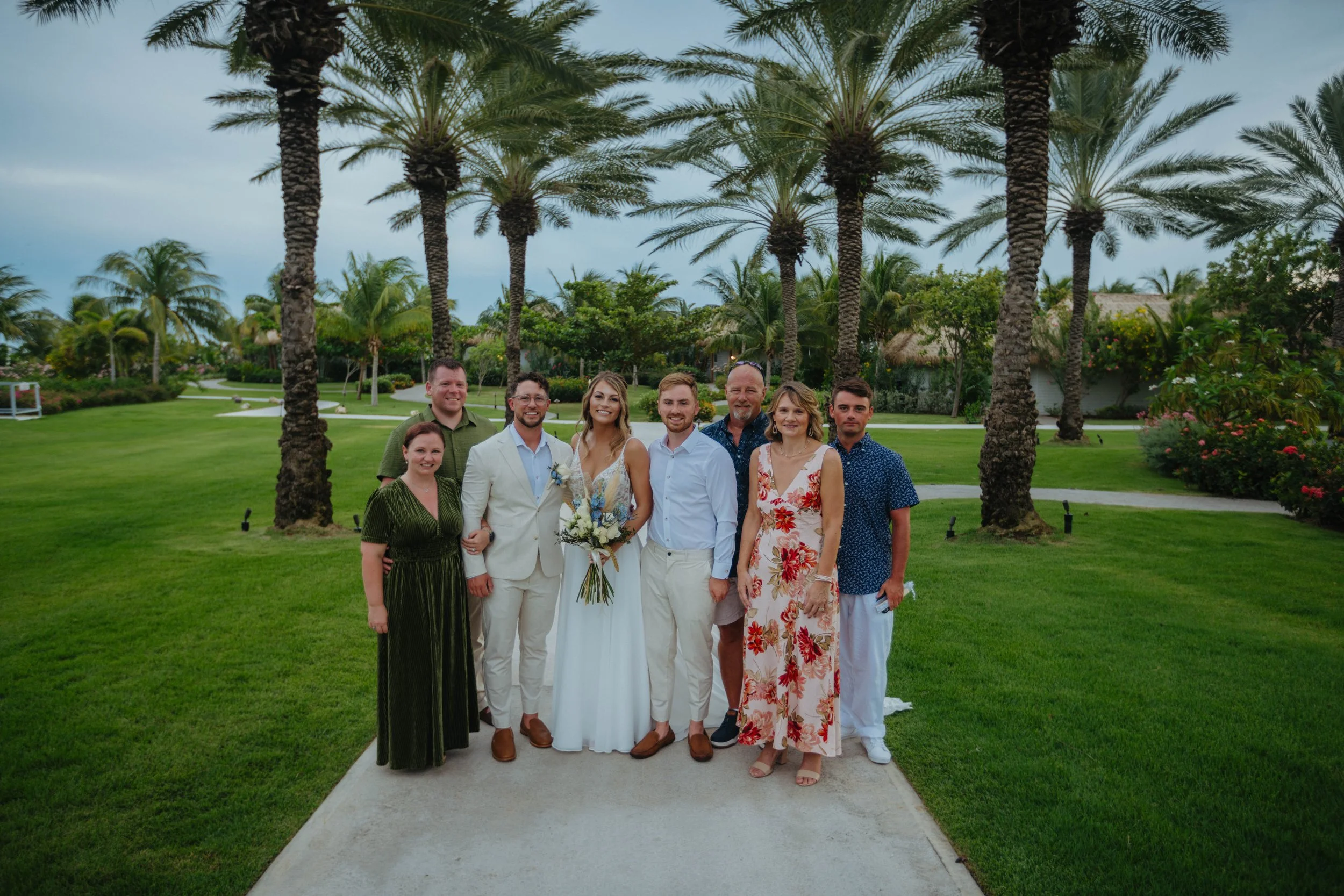 A group of nine people dressed in formal and semi-formal attire standing on a pathway in a lush green park with palm trees in the background, posing for a group photo in the daytime.