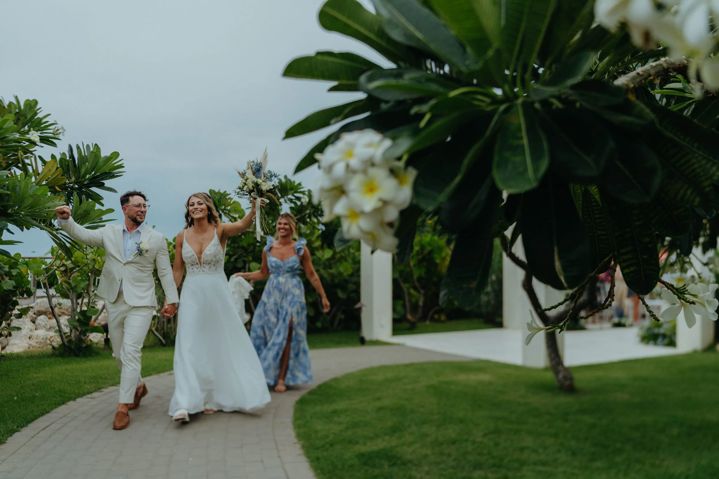 A wedding celebration with a couple holding hands and smiling, surrounded by lush green trees and plants, with a woman in a blue dress walking behind them.