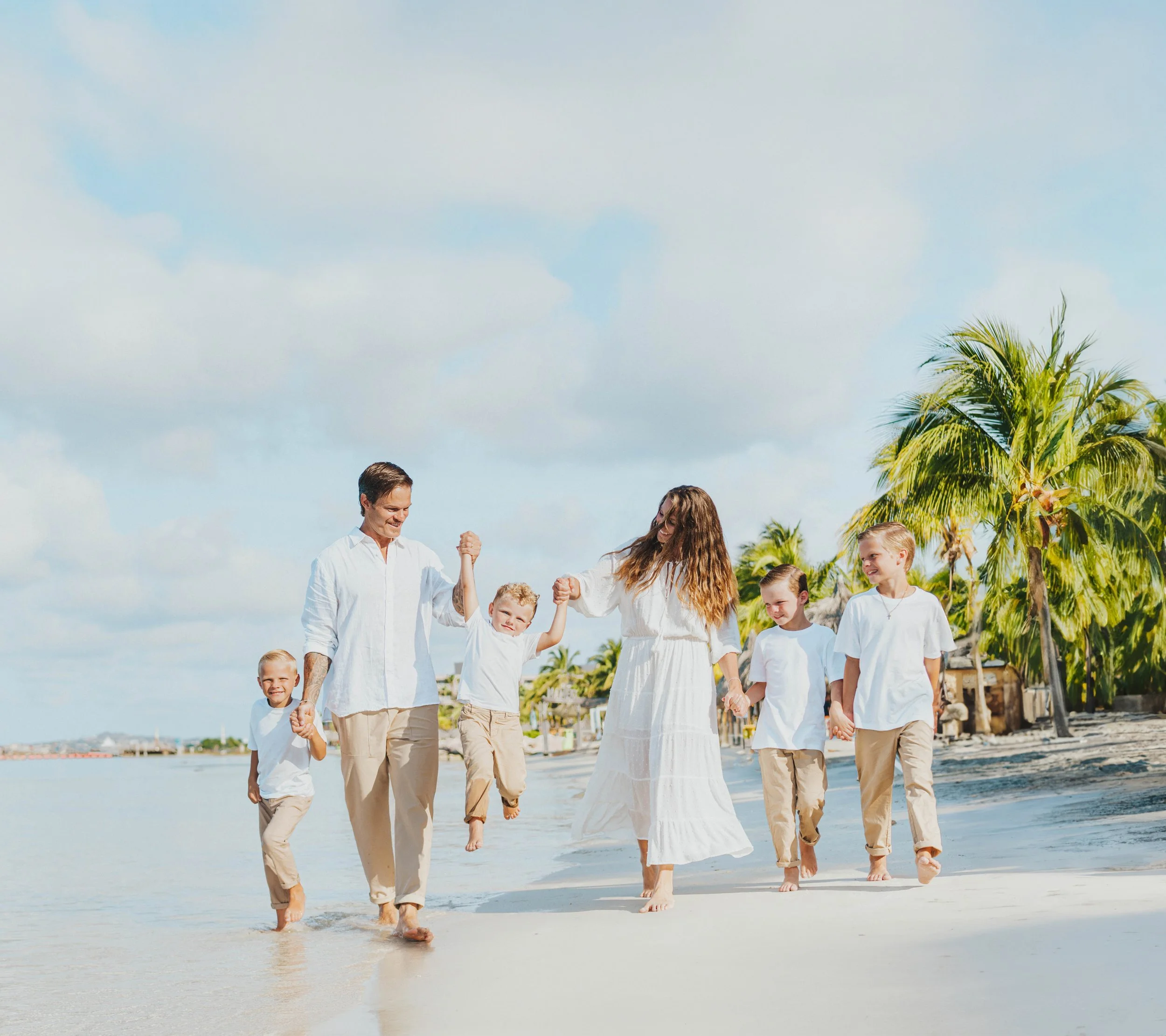 A group of people, including adults and children, walking along the beach. They are dressed in light-colored clothing, with palm trees and a cloudy sky in the background.