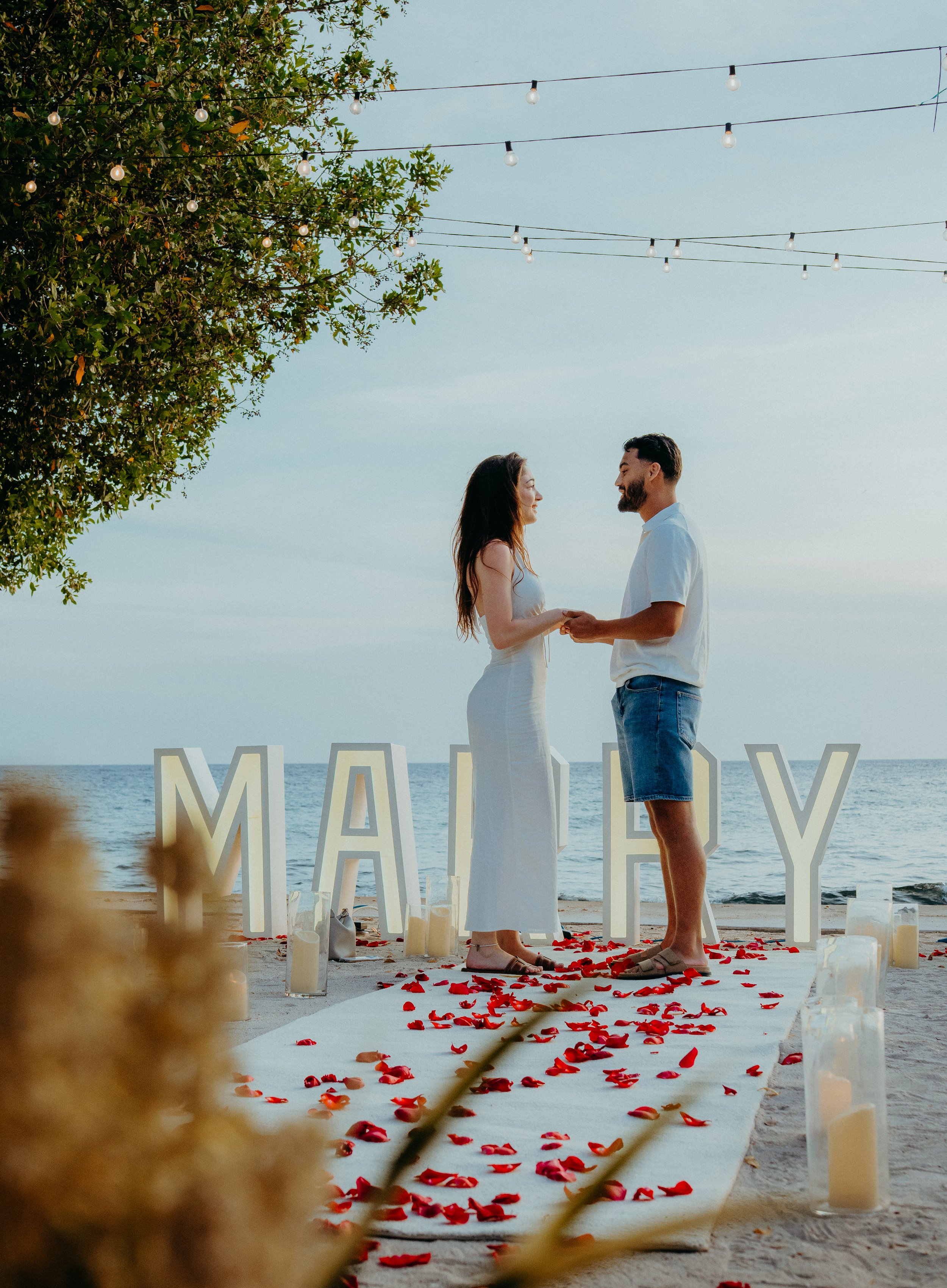 A couple holding hands during a beach wedding ceremony, with a white aisle runner decorated with red rose petals, large white letters spelling 'MARRY,' and a backdrop of the ocean and sky. They are smiling and looking at each other, under string ligh