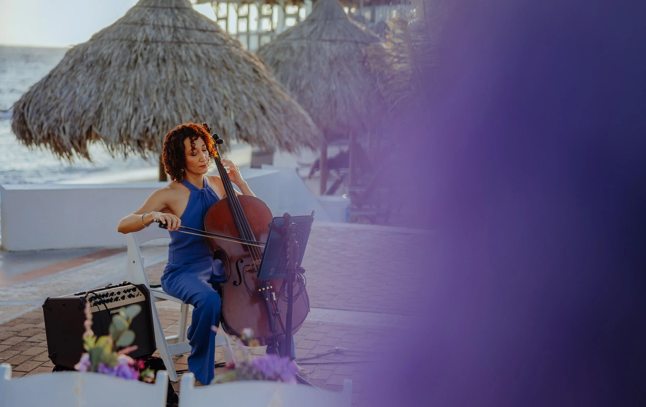 A woman with curly hair in a blue dress playing the cello outdoors near the water, with straw umbrellas and rocky shoreline in the background.