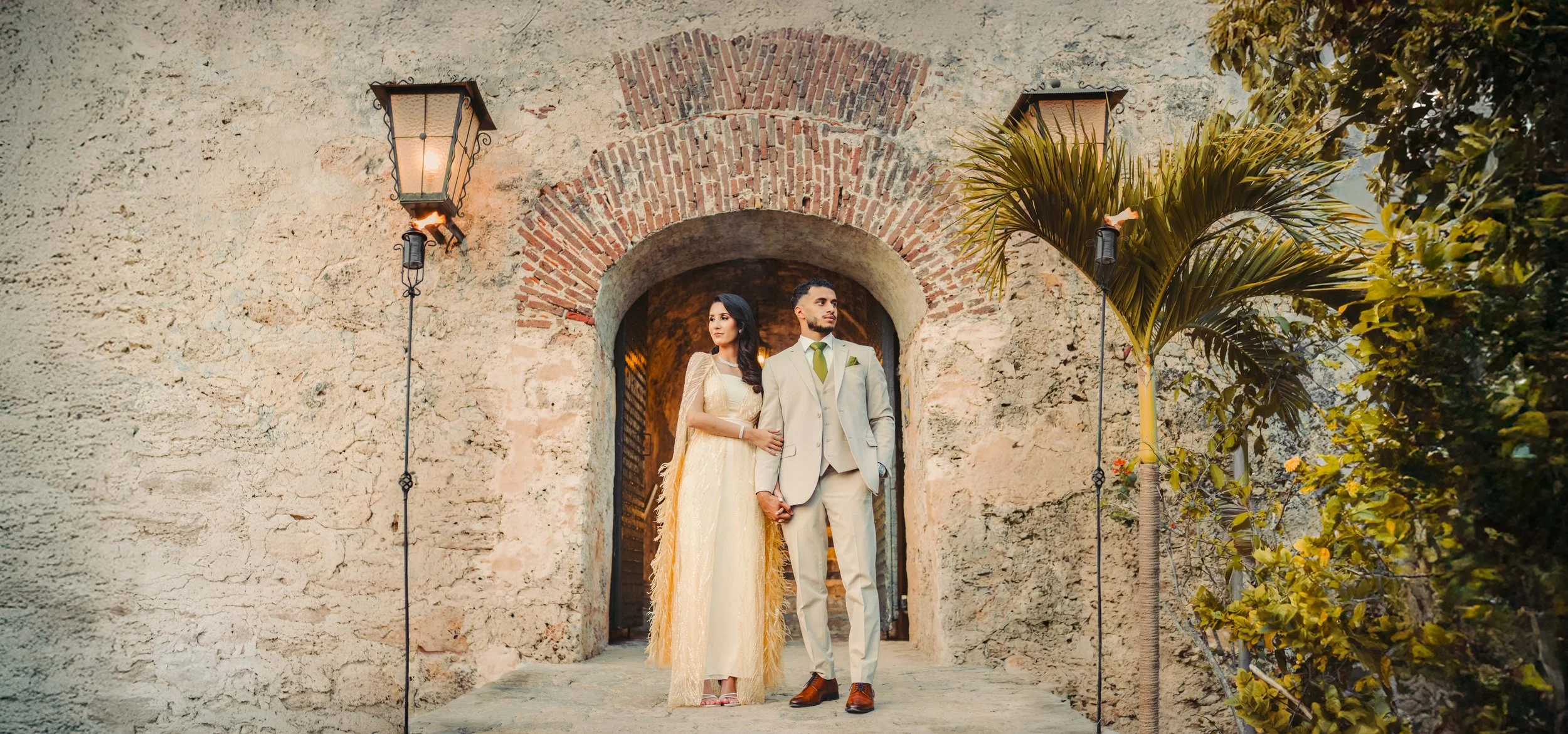 A bride and groom holding hands and standing in a stone archway outside, with trees and lights around them.