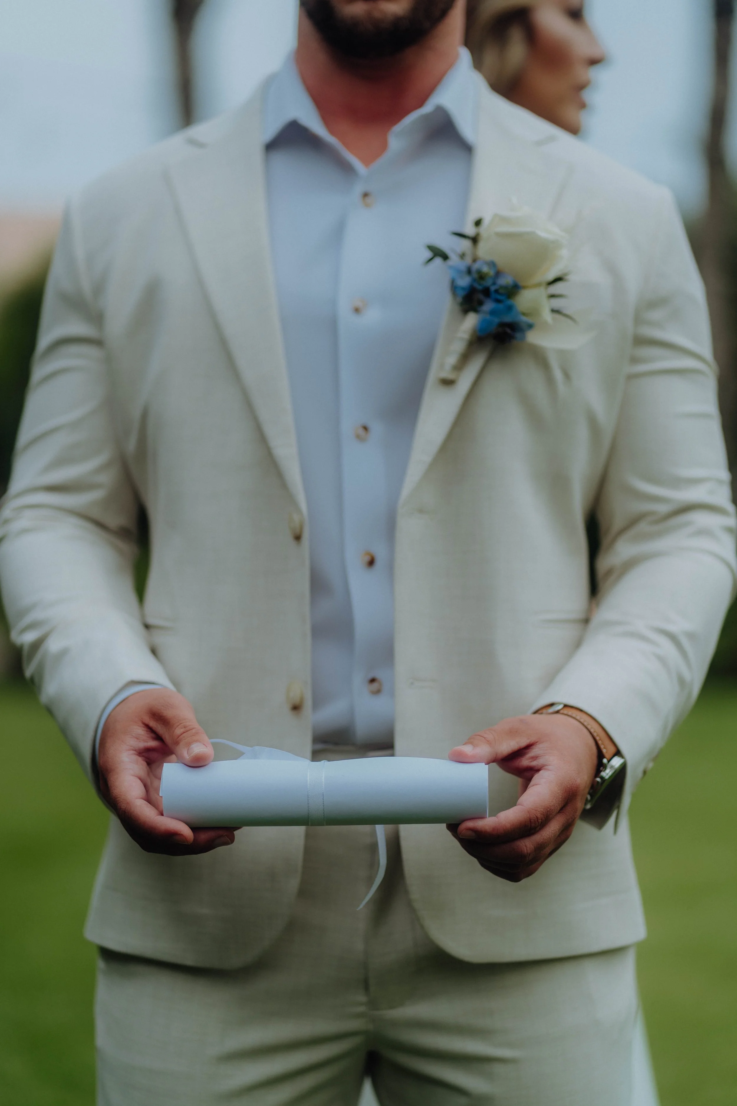 A man dressed in a light-colored suit and shirt, holding a rolled-up document, standing outdoors during a wedding or formal event. A woman in a similar suit is blurred in the background.