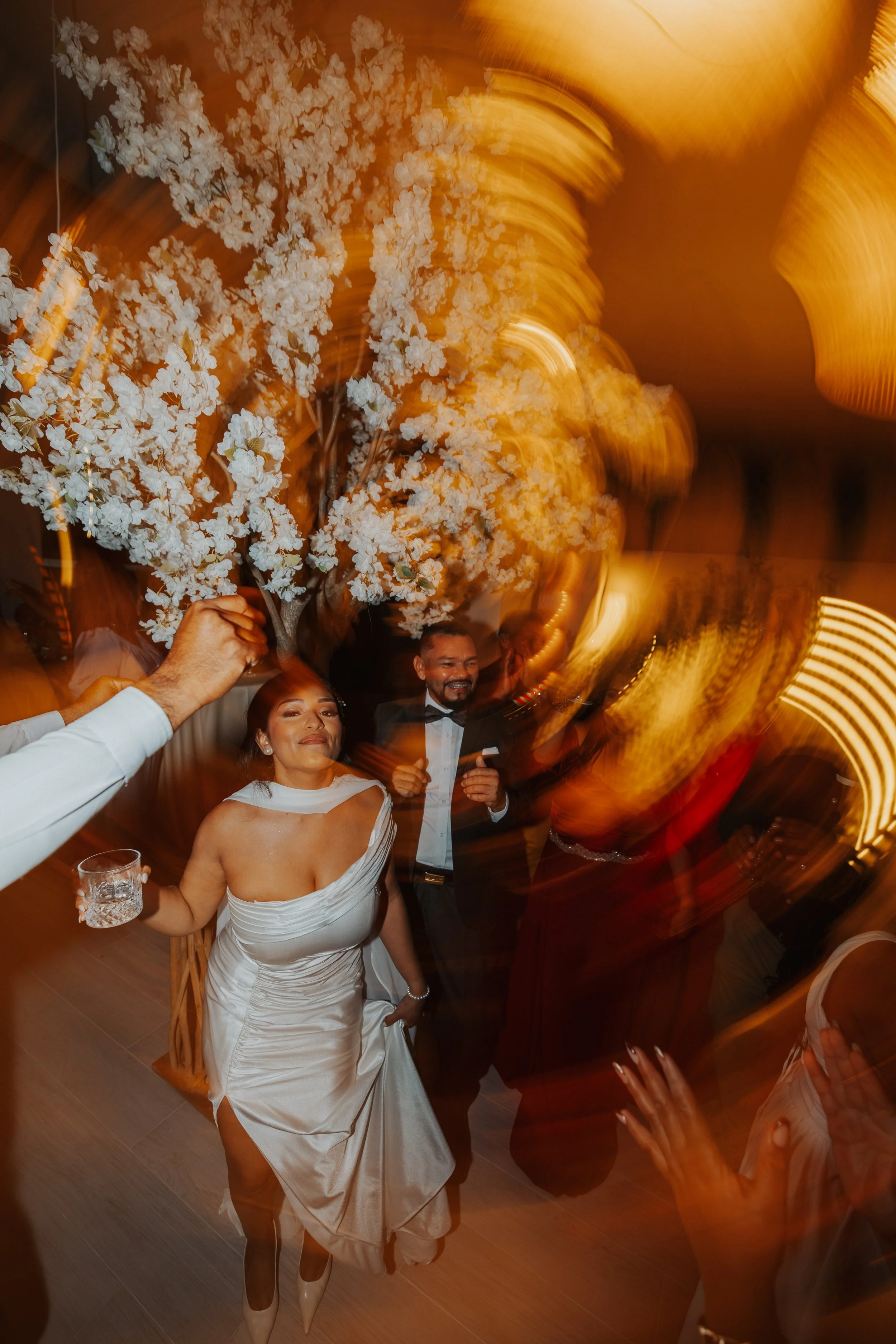 A wedding reception with a bride in a white wedding dress holding a glass, a groom in a tuxedo, and guests dancing around them under warm lighting.