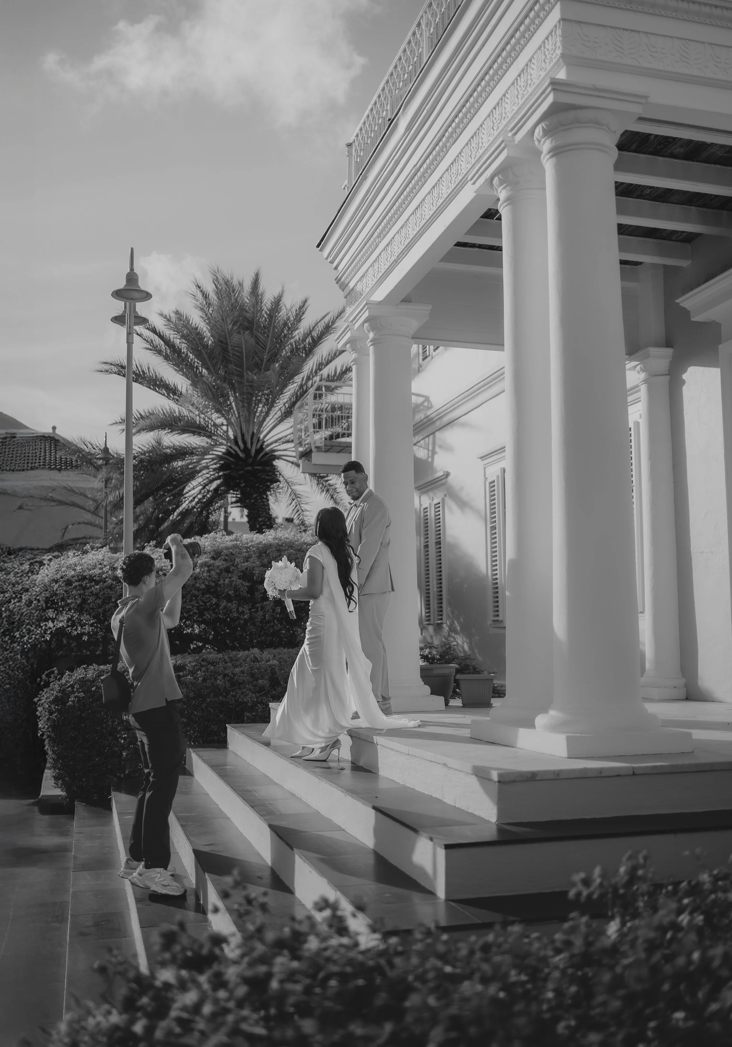 A couple is taking wedding photos outside a white, classical building with large columns and ornate trim. The bride holds a bouquet of flowers, and the groom is smiling in front of her. A person with a camera is photographing them from the ground.