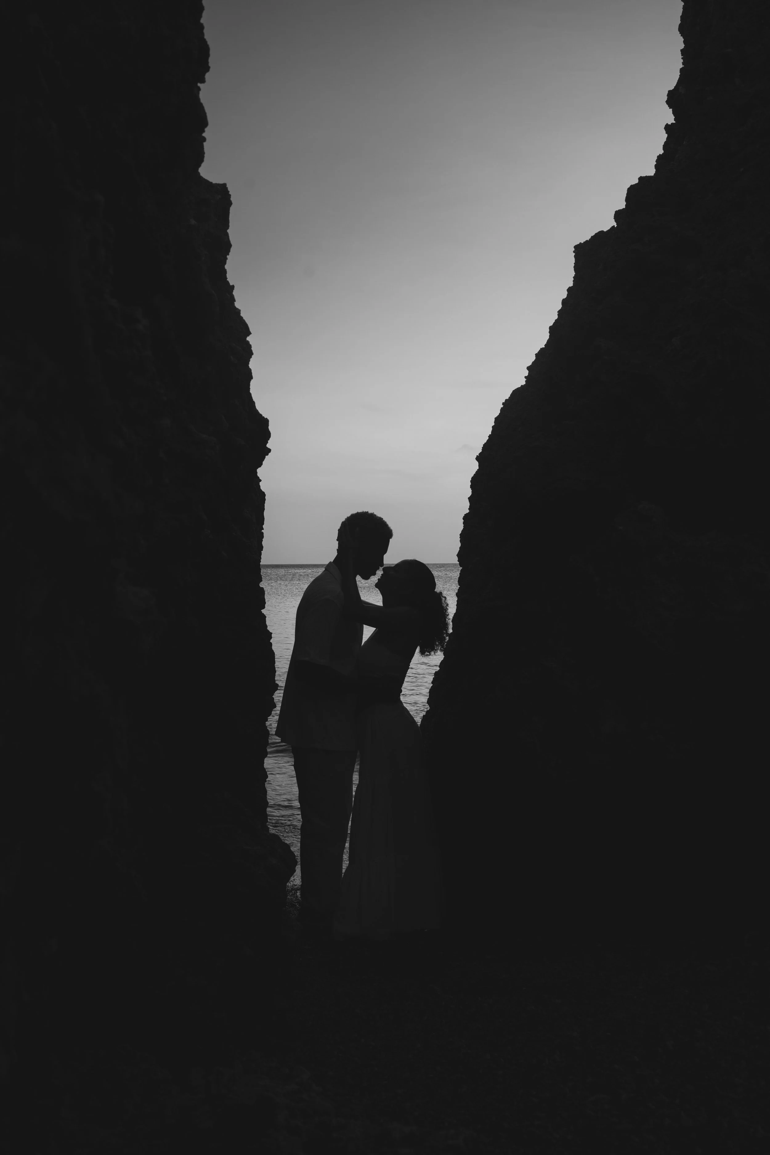 Silhouette of a couple standing close together under a rocky archway on the beach, with the sea and sky in the background during dusk or dawn.