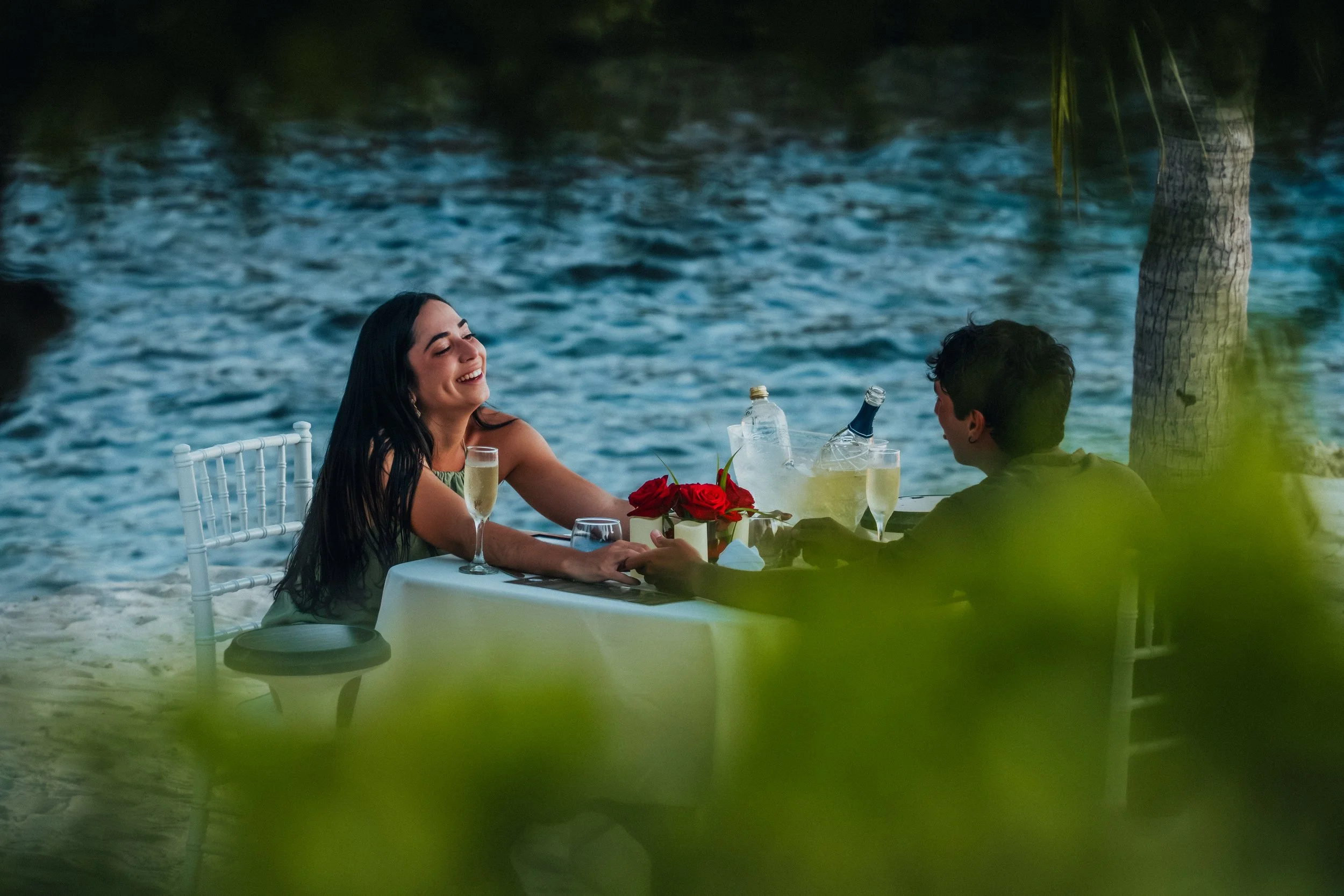 A woman and a man are sitting at a table outdoors near water, enjoying a meal with drinks and a bouquet of red roses.
