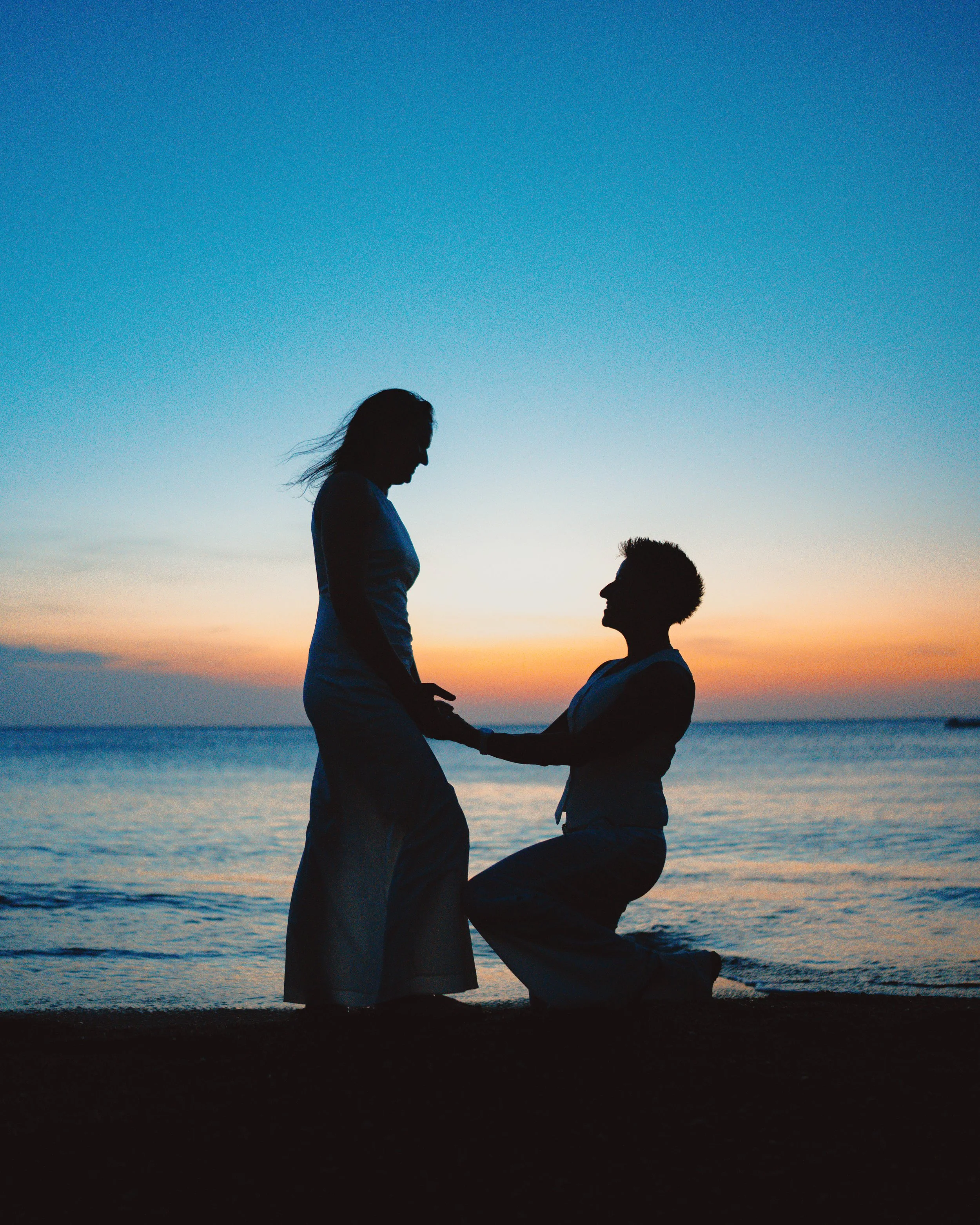 Silhouettes of a woman and a boy at the beach during sunset, holding hands, with the ocean and a colorful sky in the background.