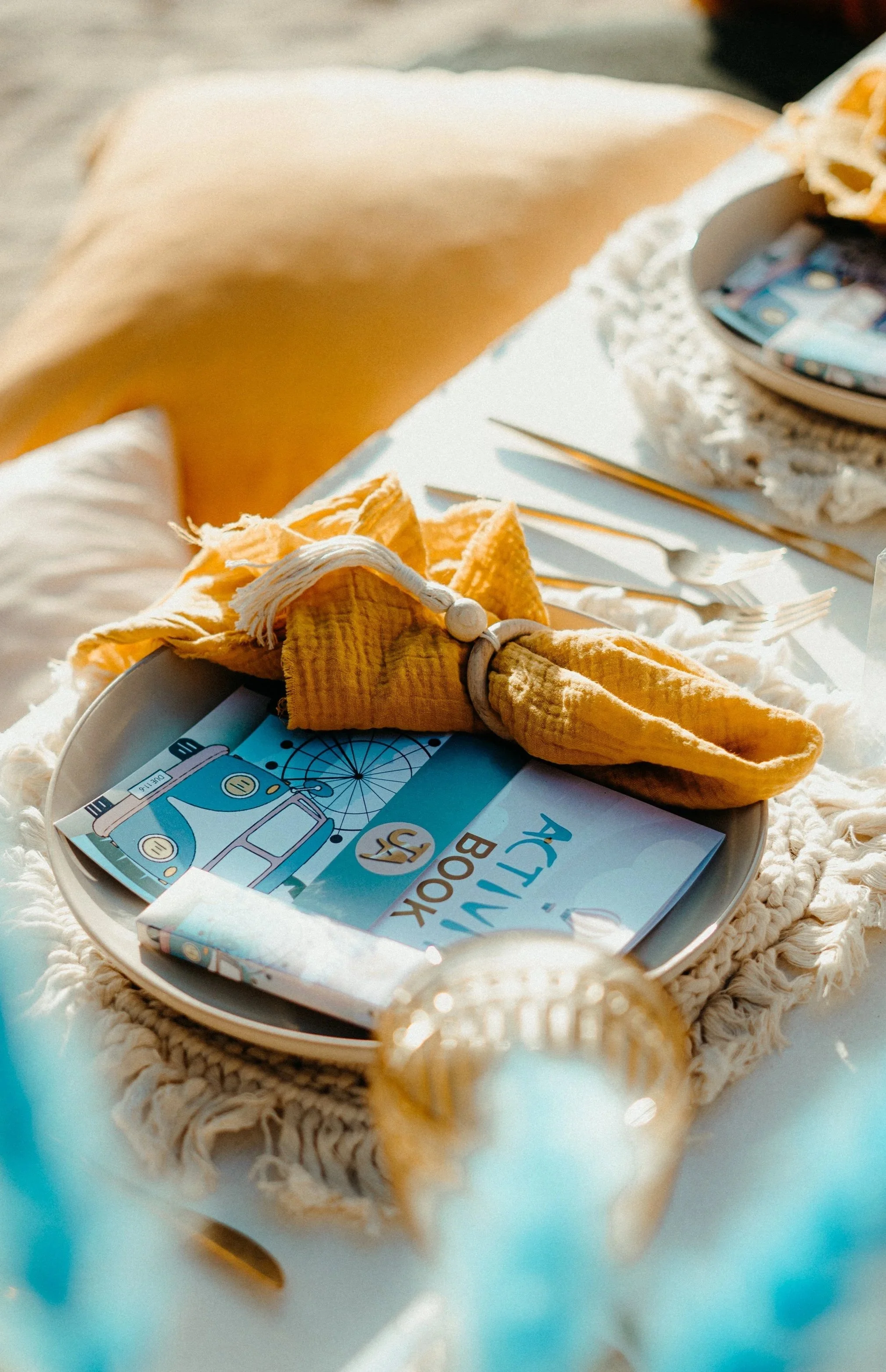 A round tray with a blue, yellow, and white book titled 'ACTIVITY BOOK,' a folded yellow cloth, and some yellow fabric with white tassels. The tray is on a white layered cloth.