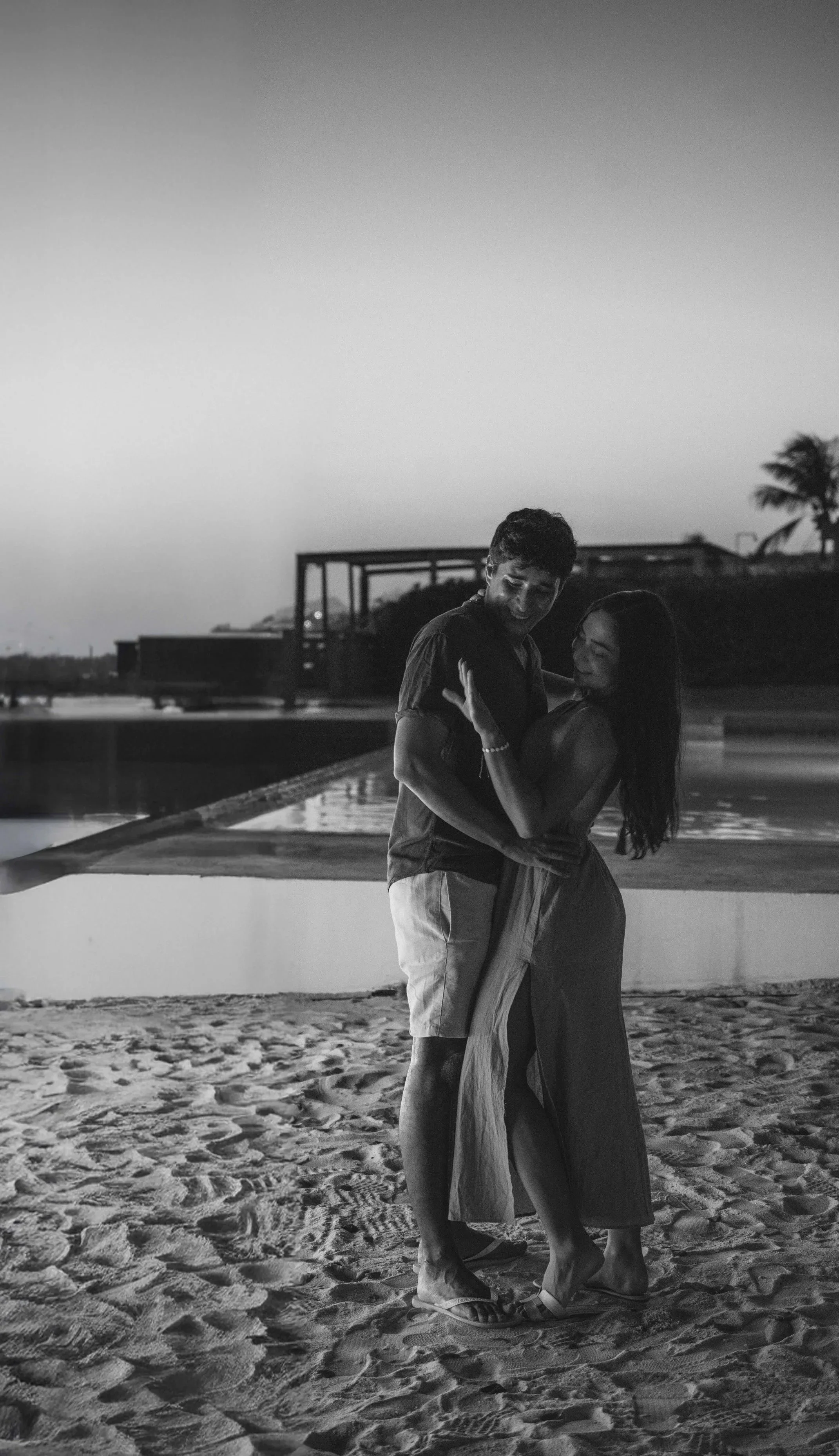 A black and white photo of a couple dancing on a sandy beach during sunset or twilight, with the ocean and some palm trees in the background.
