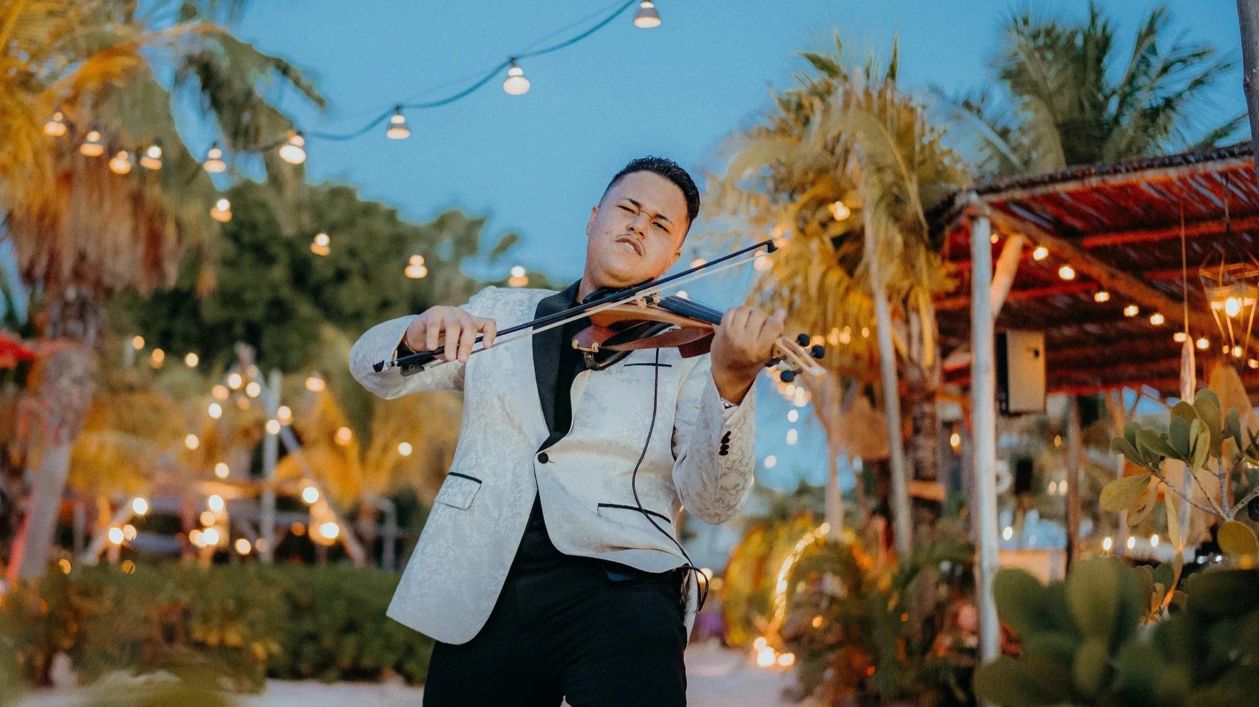 A man in a white patterned suit jacket playing the violin at an outdoor event during dusk, with string lights and trees in the background.