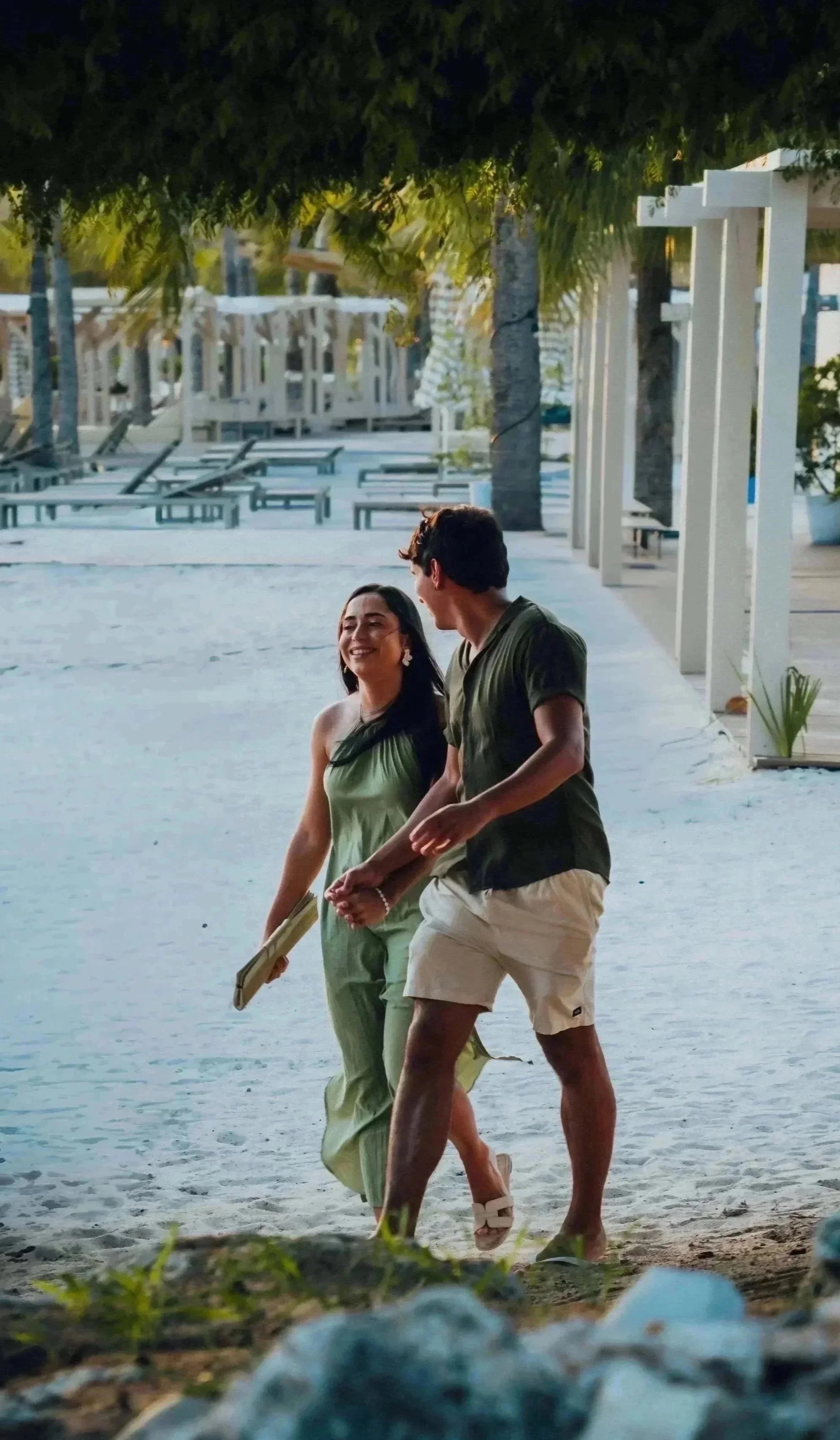 A young man and woman walking along a beach shore at sunset, smiling and talking, with palm trees and beach chairs in the background.