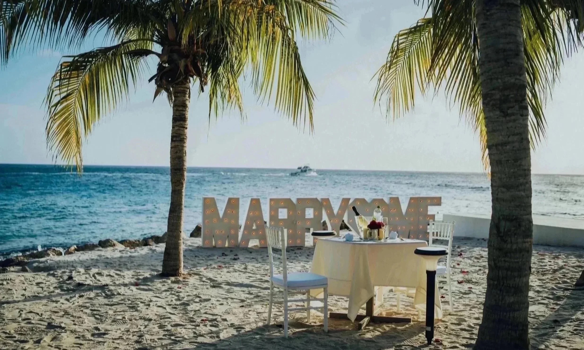 Beachside dining setup with a table and chairs, palm trees, a large illuminated "XOXO" sign, ocean waves, and a boat in the distance under a clear sky.