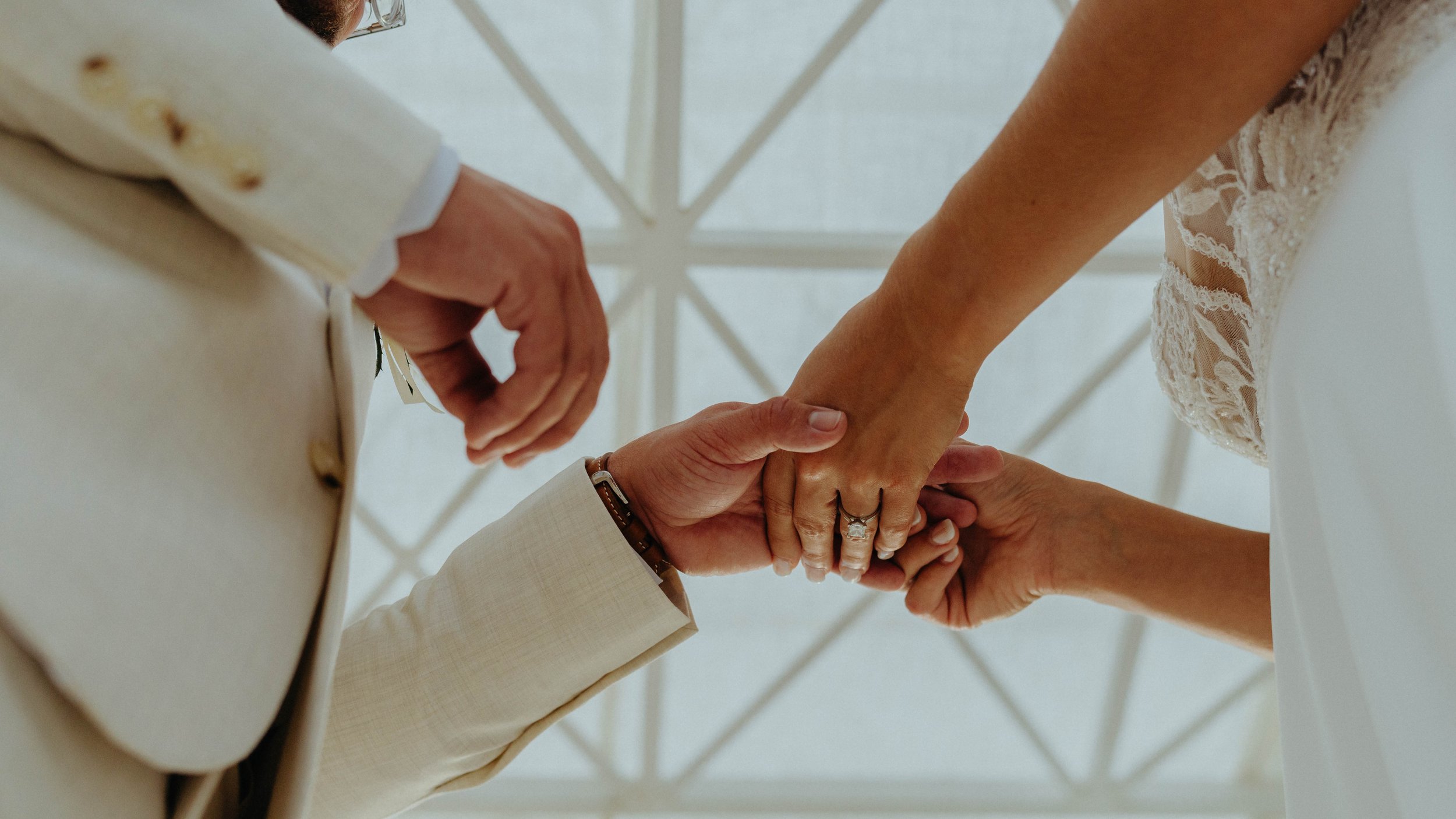 Close-up of a couple's hands holding each other, with the wedding rings, seen from below against a background of a white canopy structure.