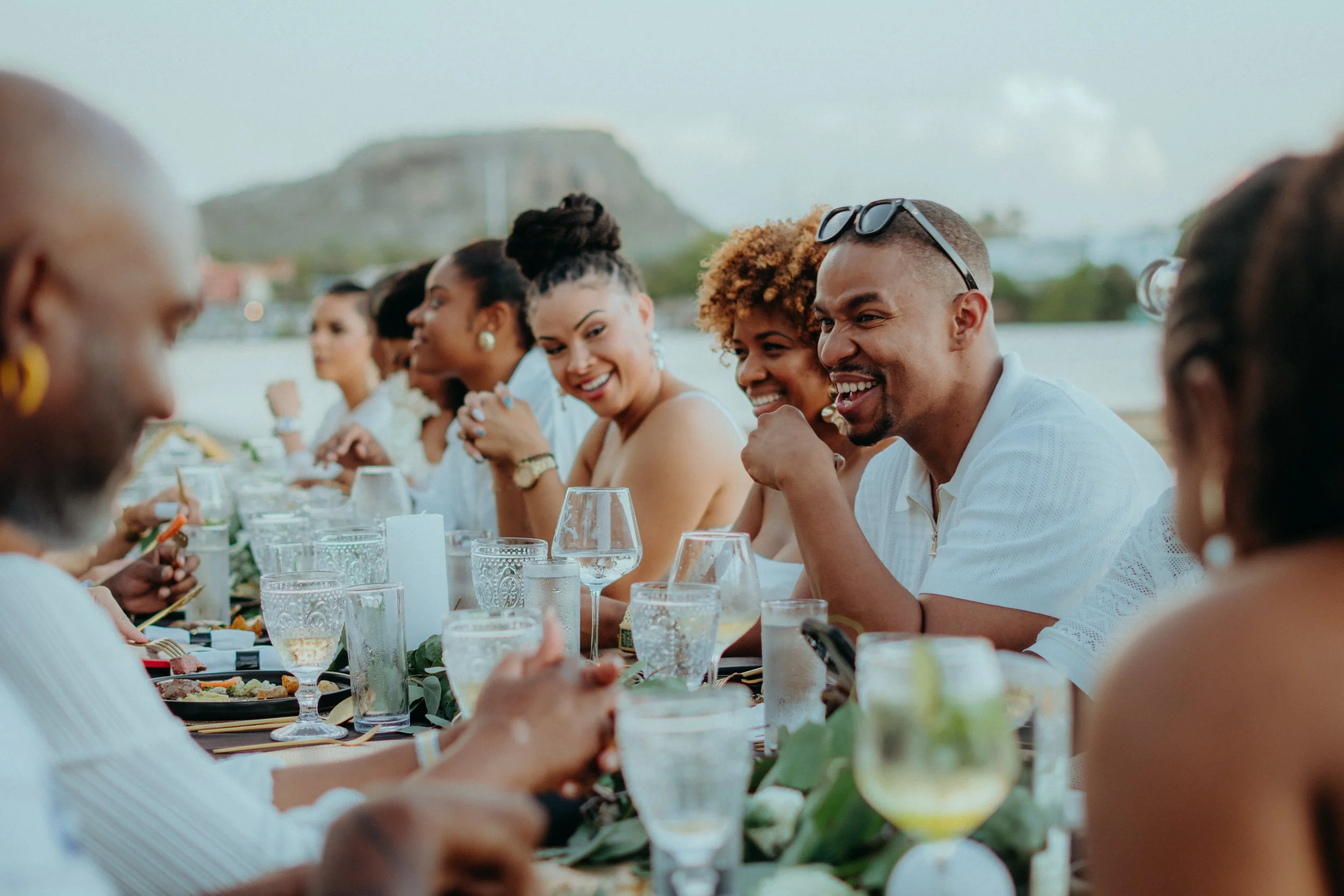 People sitting at a long outdoor dining table, enjoying food and drinks, with a view of a mountain and water in the background.