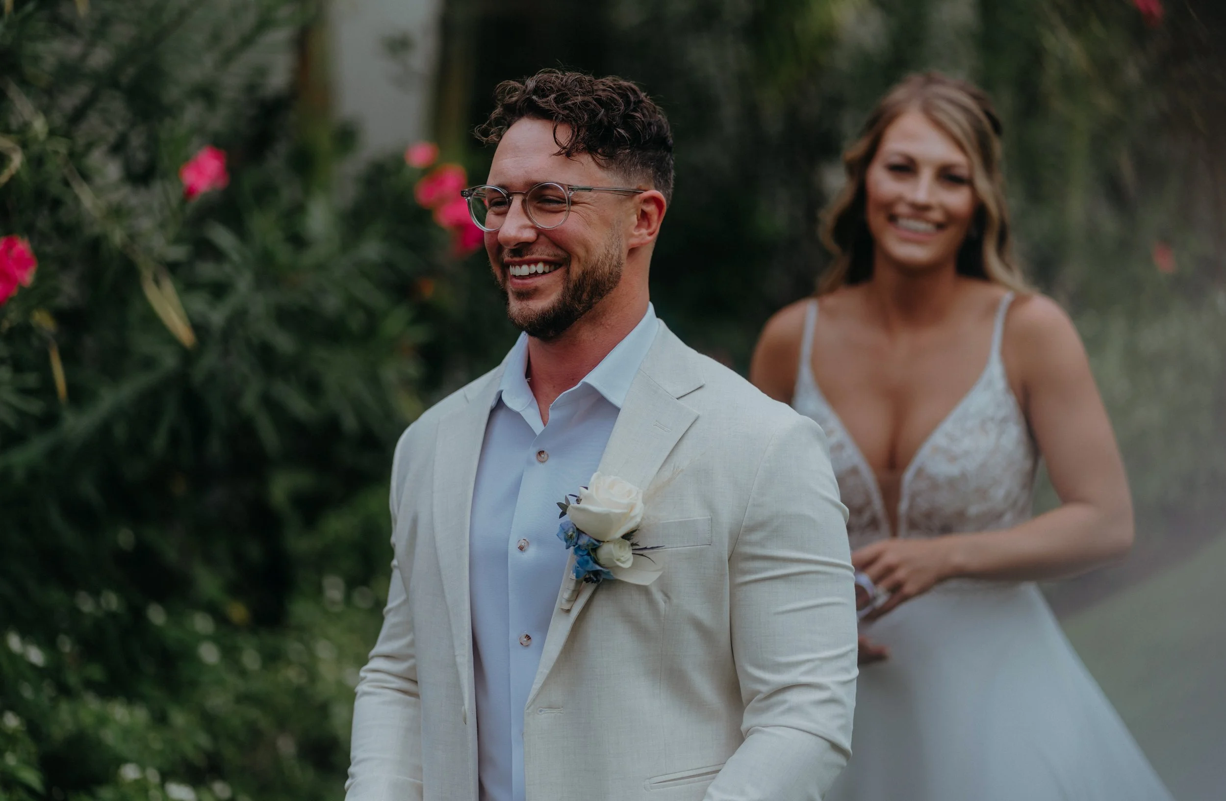 A smiling man in a light suit with a boutonniere, and a woman in a wedding dress, walking outdoors among green bushes and pink flowers.