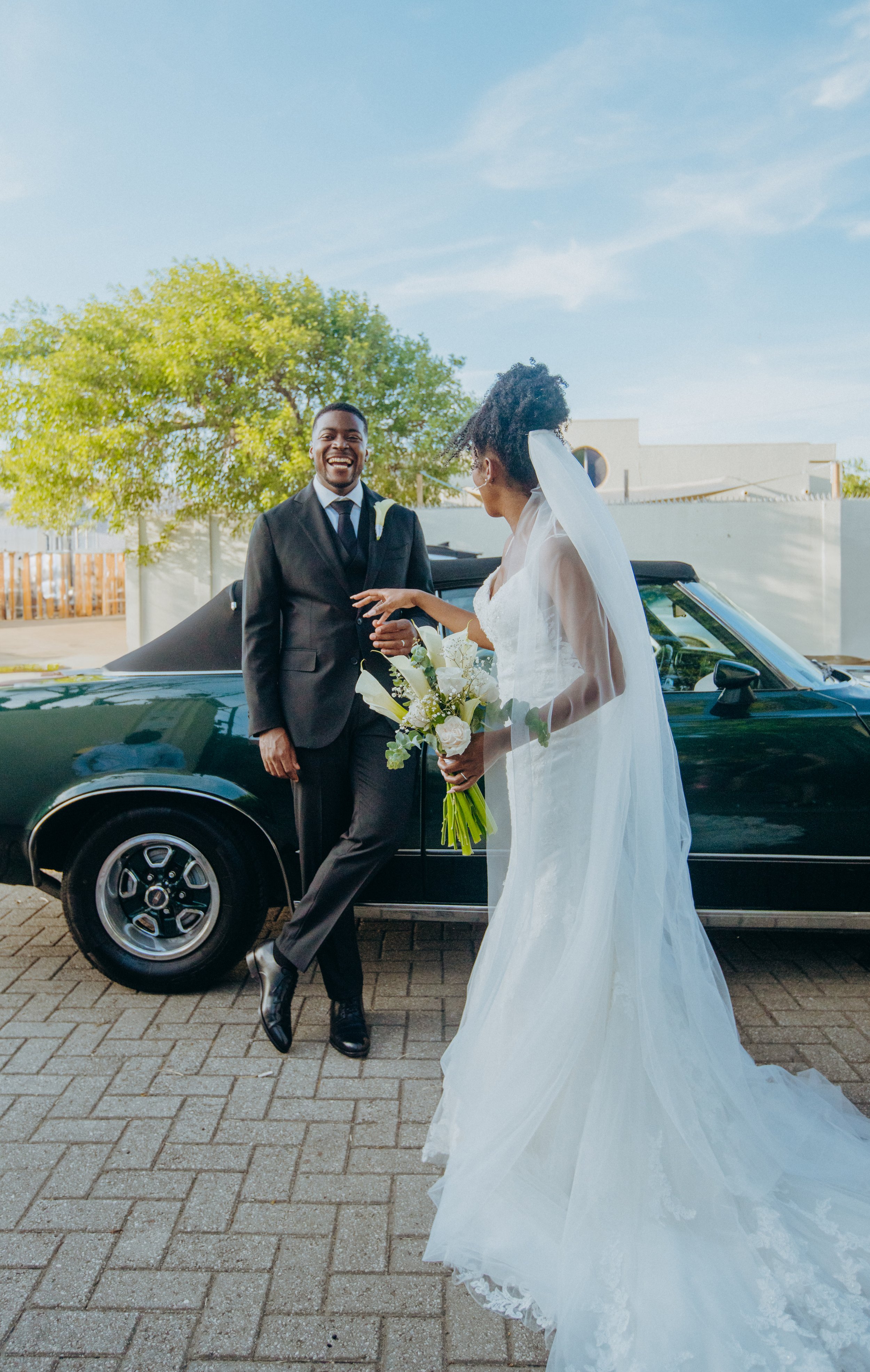 A newly married couple standing near a black vintage car outdoors, with the groom in a black suit and the bride in a white wedding dress holding a bouquet, smiling and facing each other under a clear blue sky and green trees.