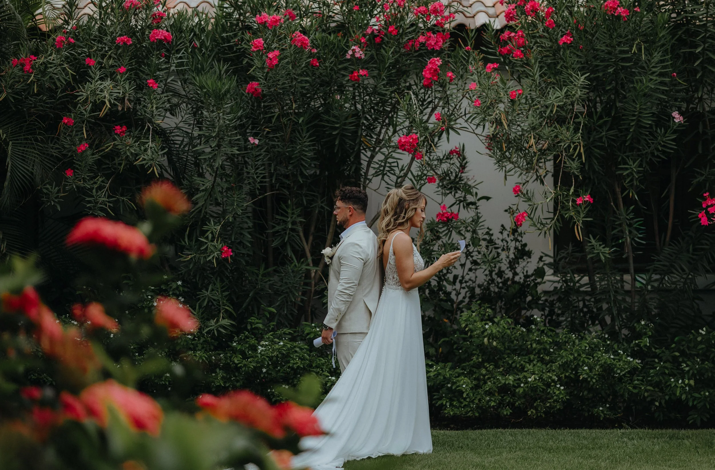 A bride and groom standing together outdoors in front of tall green bushes with pink flowers, the bride holding a piece of paper and both dressed in wedding attire.