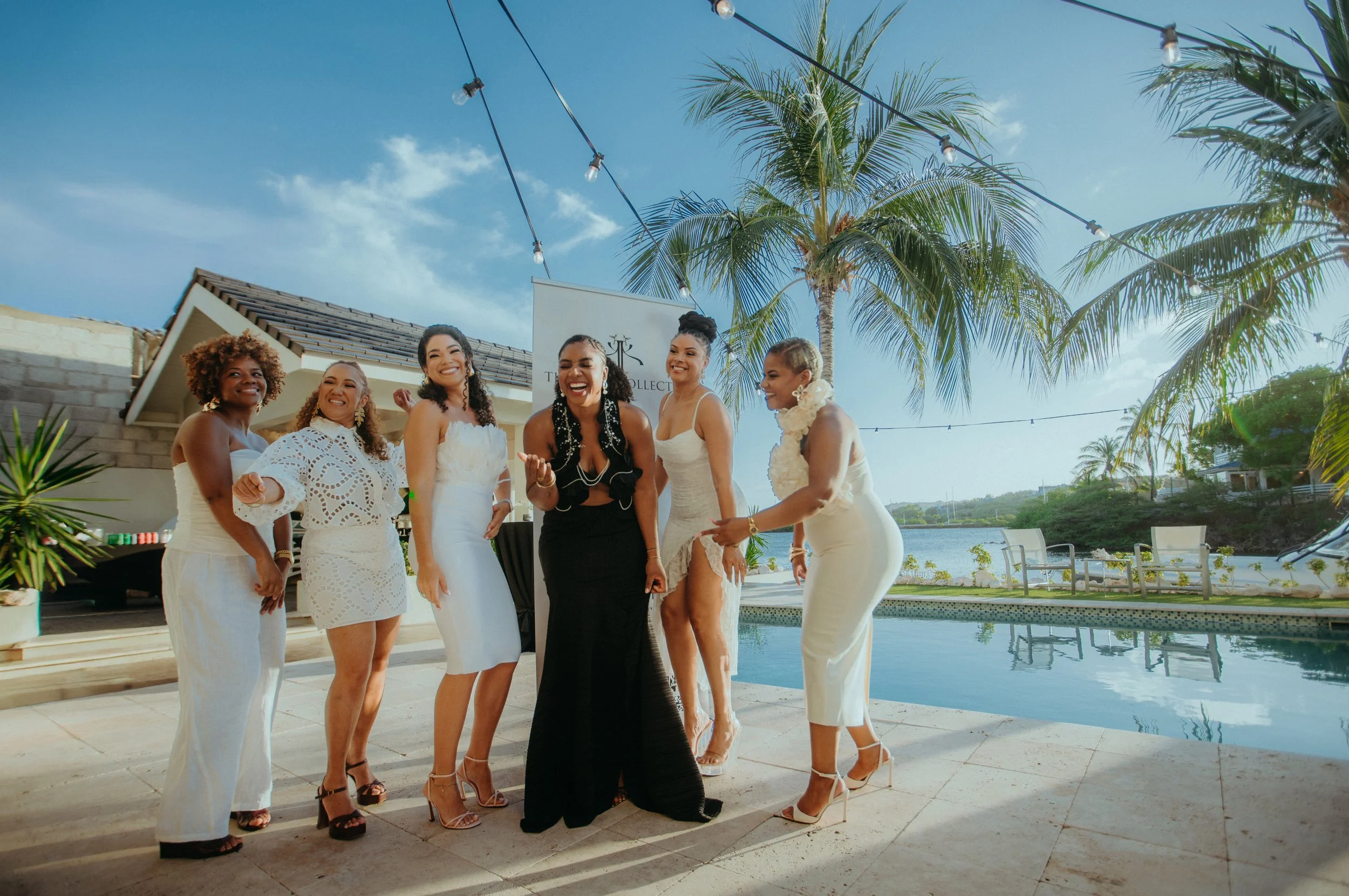 Group of women in white dresses and black dress smiling and dancing by a poolside under string lights and palm trees, with a house and lake in the background during daytime.