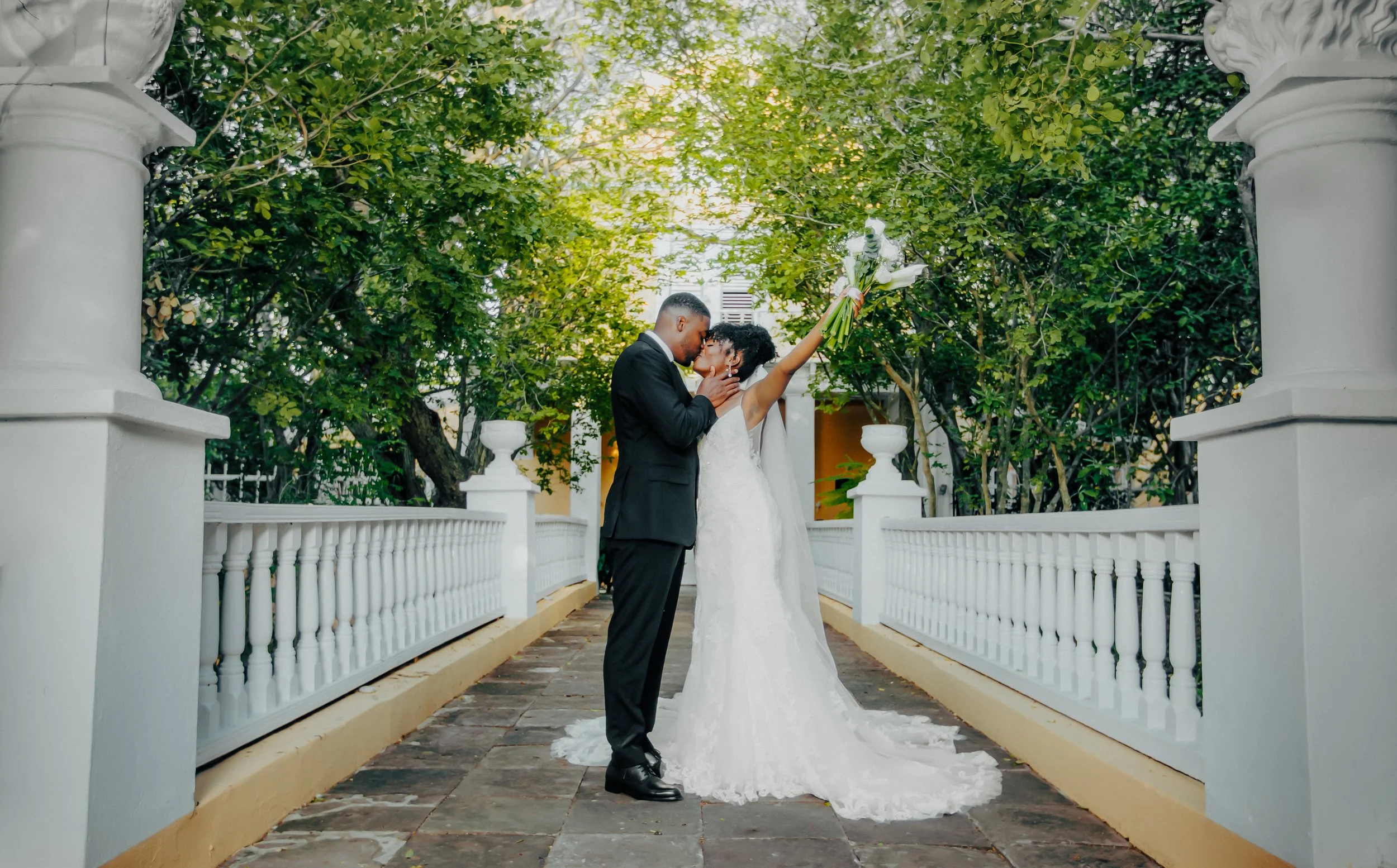 A couple in wedding attire sharing a kiss under a canopy of green trees on an outdoor deck with white railing, with the bride holding a bouquet of flowers raised in the air.