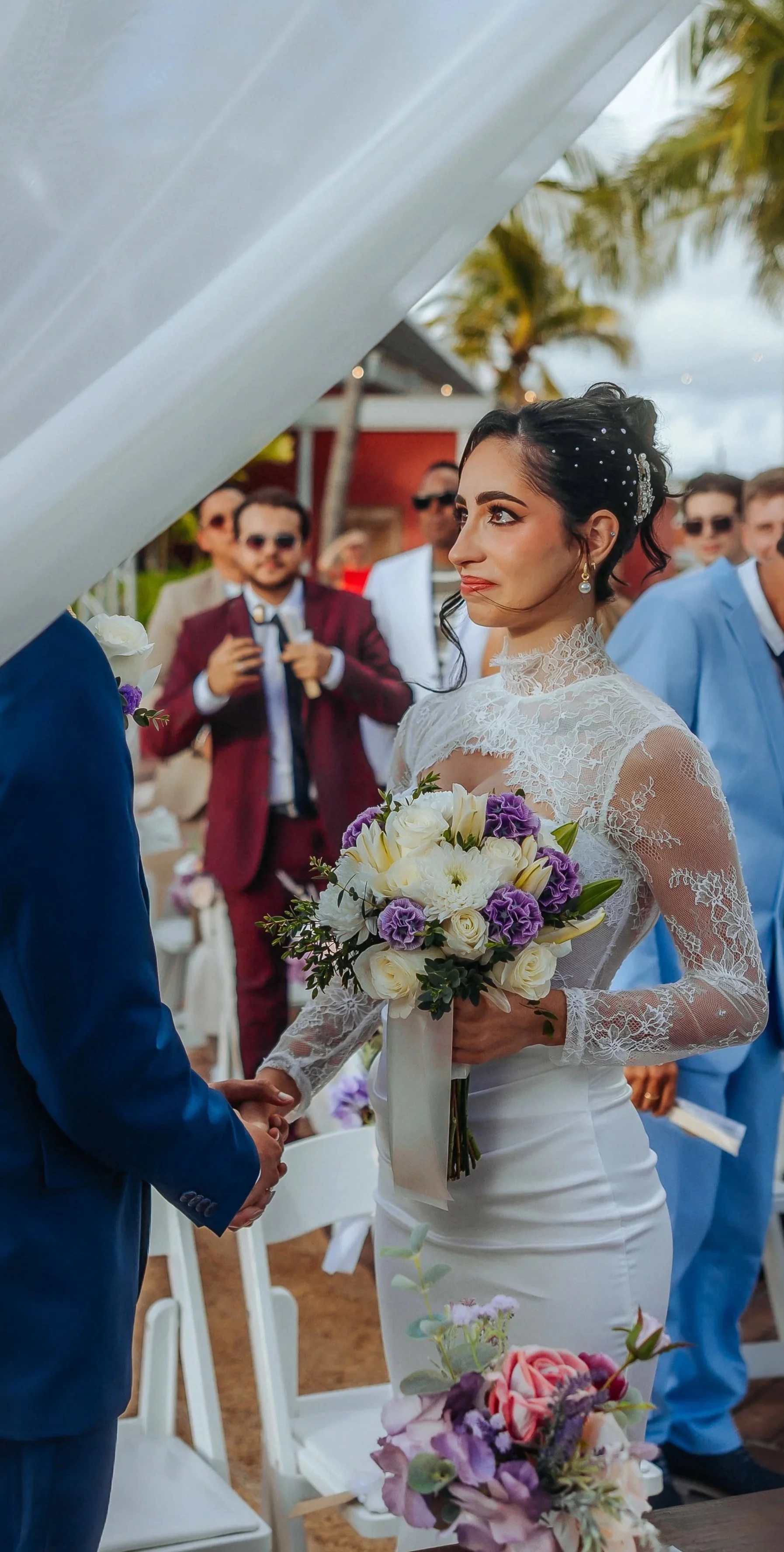 A bride and groom holding hands during an outdoor wedding ceremony with guests in the background. The bride is wearing a lace wedding dress and holding a bouquet of white and purple flowers. The groom is in a blue suit. Palm trees and a red building are visible in the background.