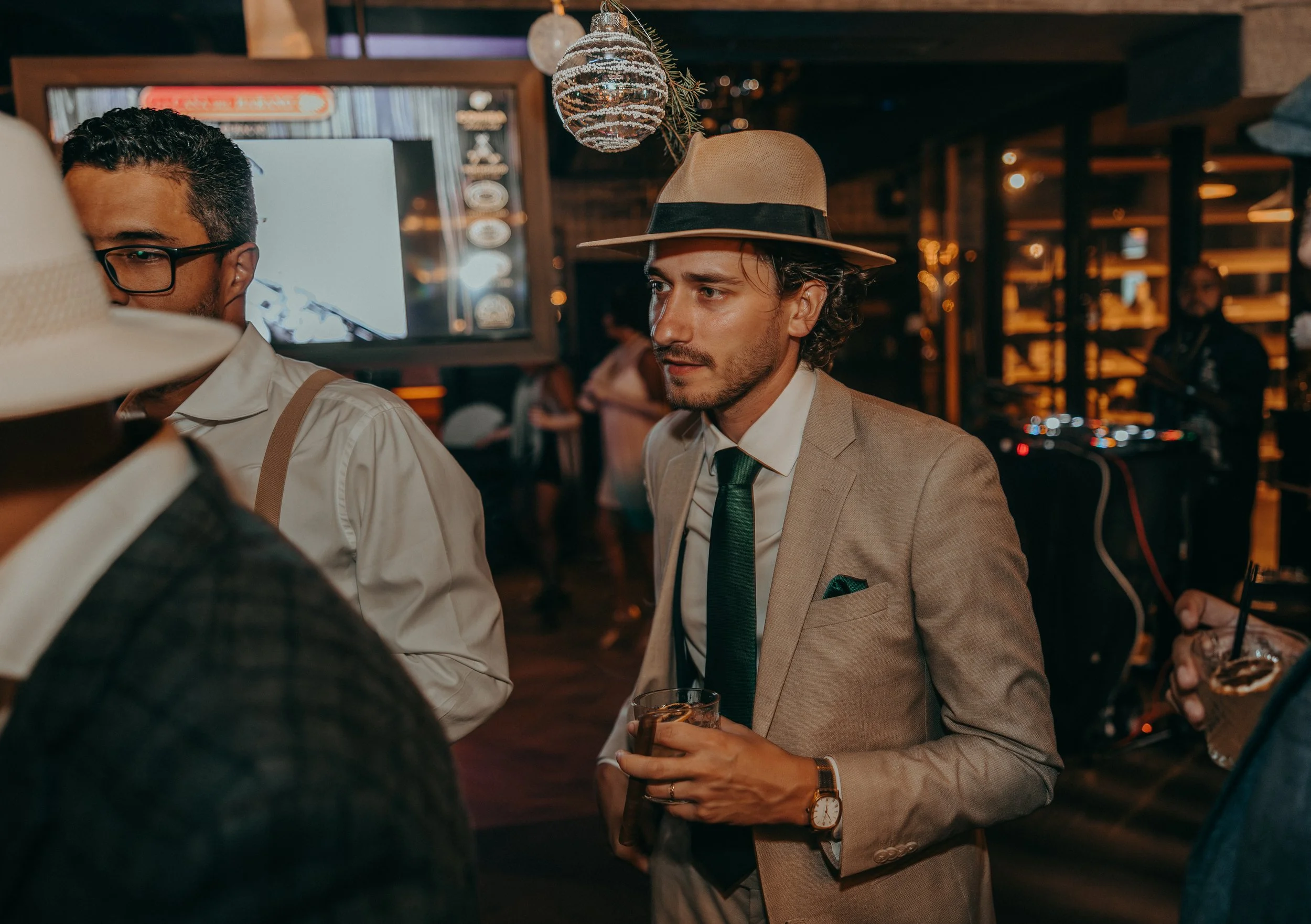 A man in a beige suit and tie holds a drink and wears a wide-brimmed fedora with a black band, at a dimly lit bar or restaurant with a rustic decor and a decorative hanging orb light.