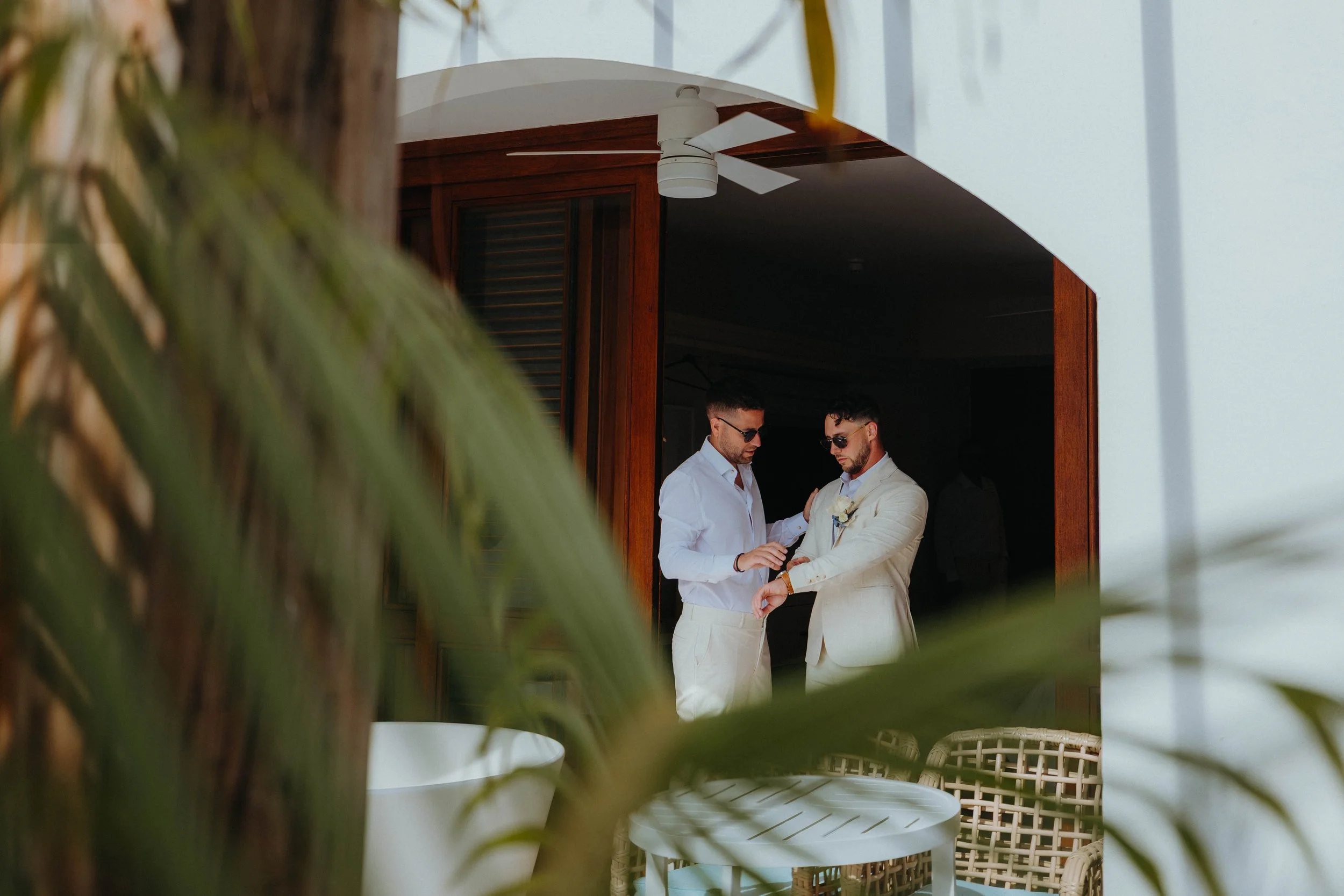 Two men in white suits and sunglasses are changing a wristwatch. They are standing outside a building with wooden window shutters and a ceiling fan. The photo is framed by blurred green plants in the foreground.