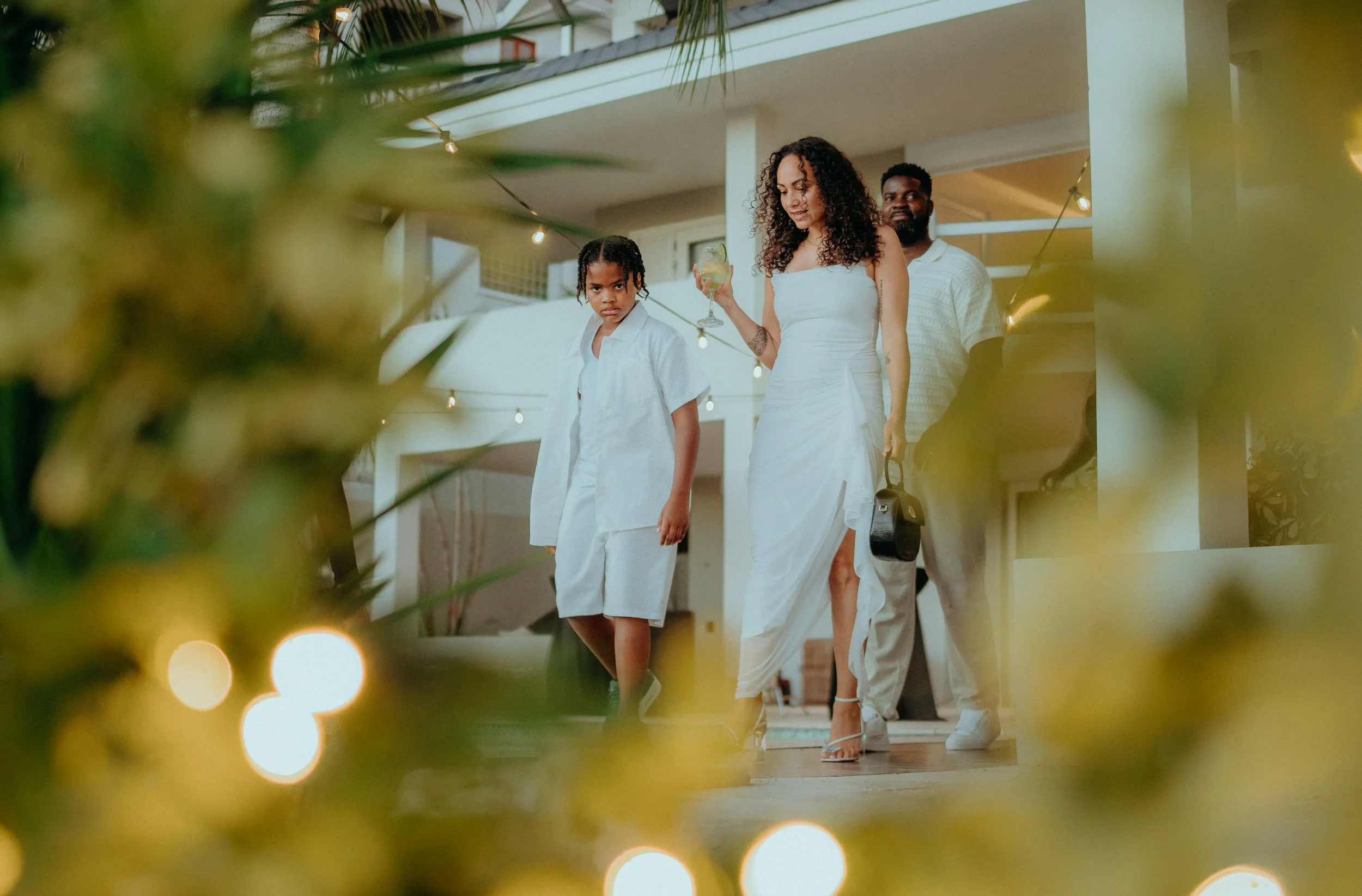 Four people, two women and two men, are walking inside a house, viewed through foliage in the foreground. The woman in front is wearing a white dress and high heels, holding a drink. The other woman is also in white, with wet curly hair, and has a se