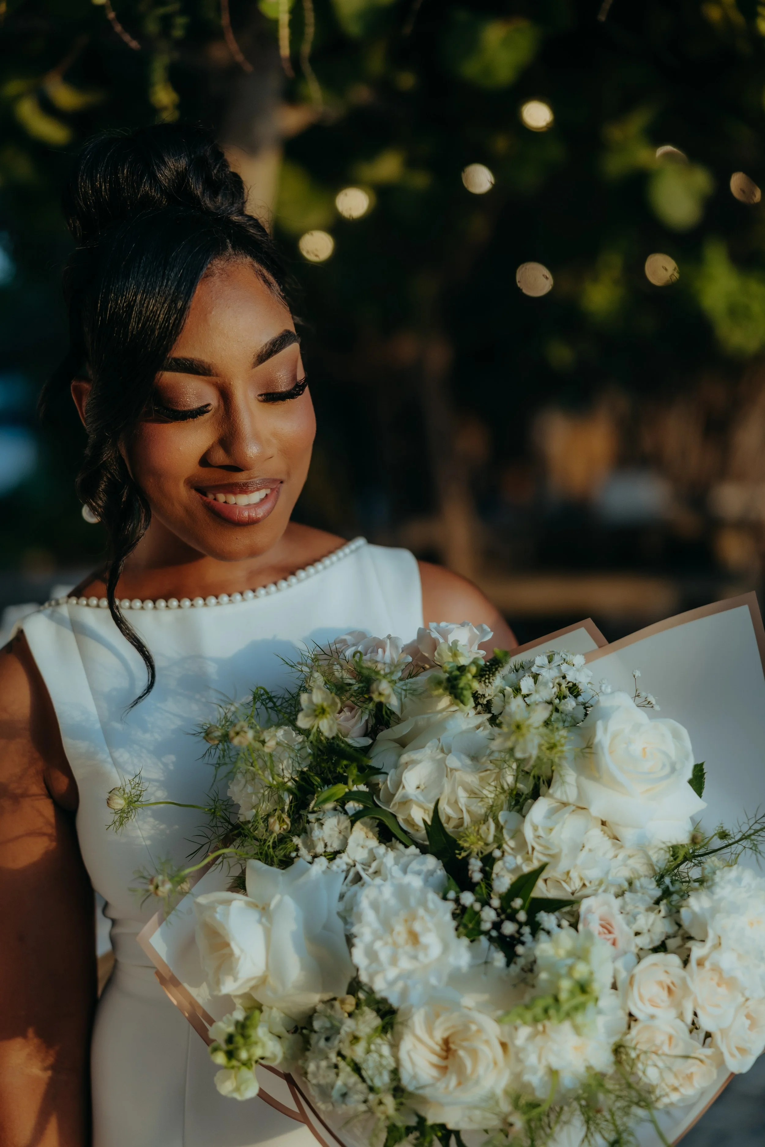 A bride with dark hair styled in an updo, wearing a white dress with pearl-embellished neckline, is smiling while holding a large bouquet of white and light pink flowers.