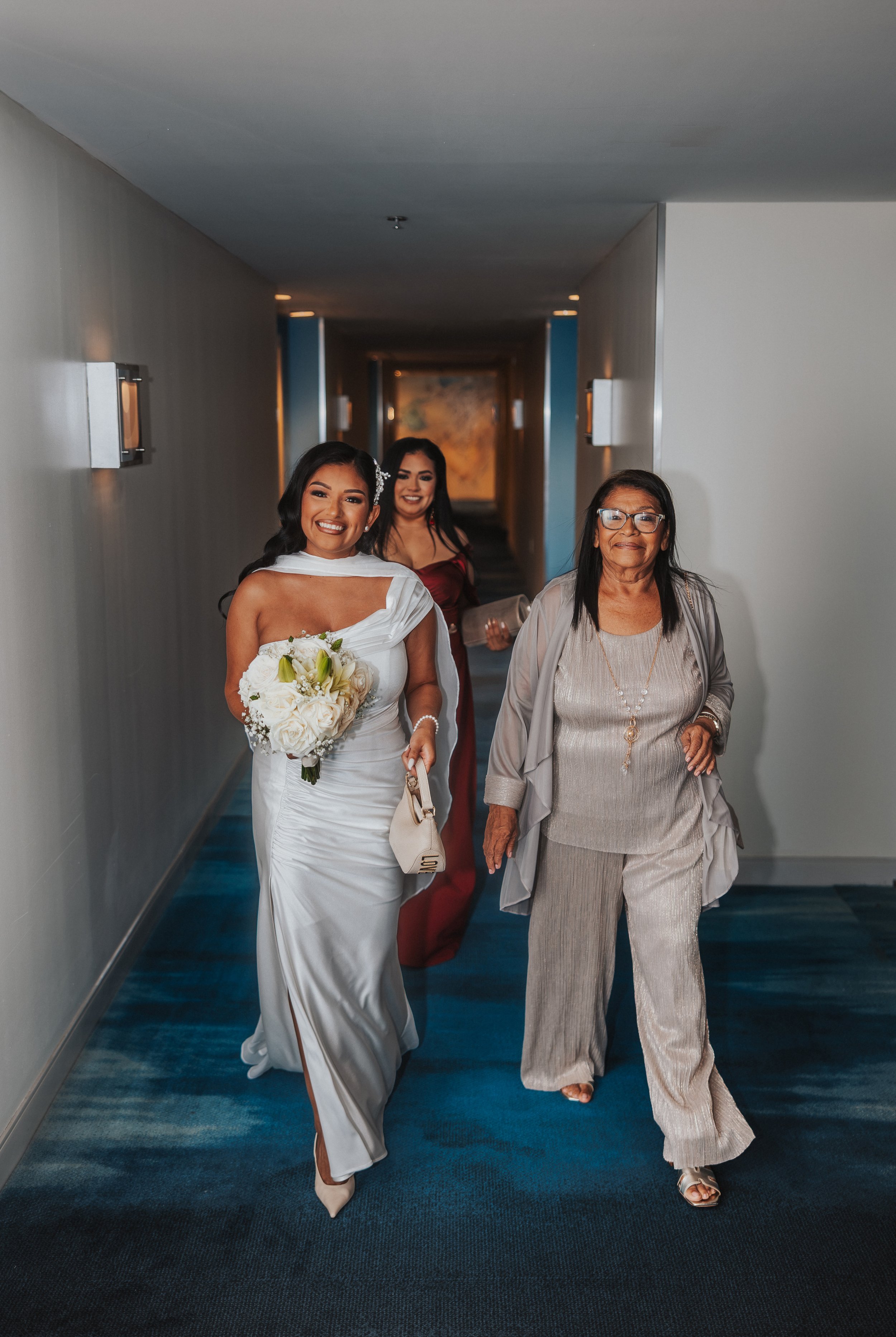 A bride in a white wedding dress holding a bouquet, walking down a hallway with two women, one in a champagne-colored outfit and one in a maroon dress, smiling as they walk together.