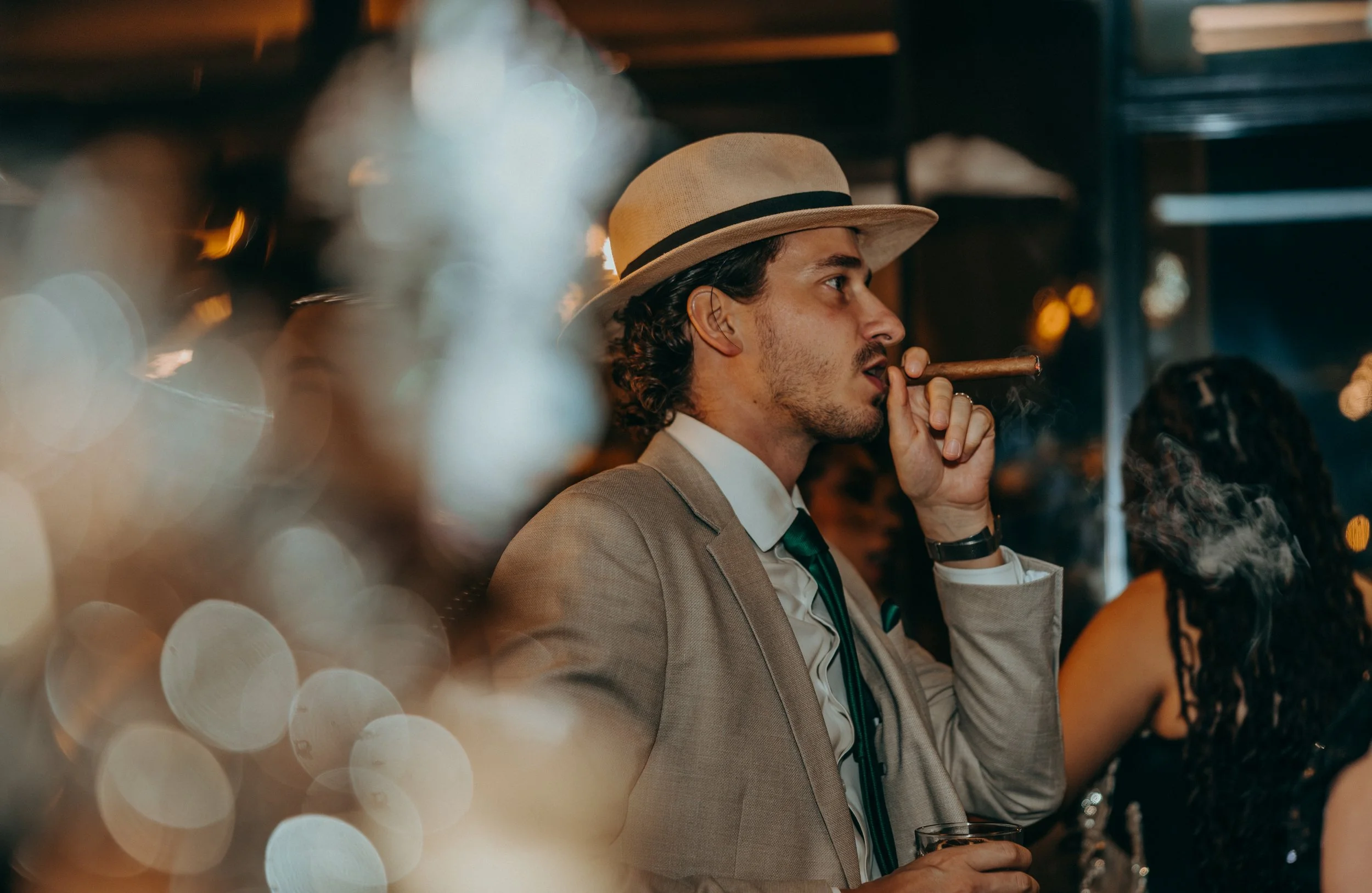 A man in a tan suit and green tie, wearing a beige fedora, smokes a cigar at a social event in a dimly lit setting with blurred lights and other attendees.