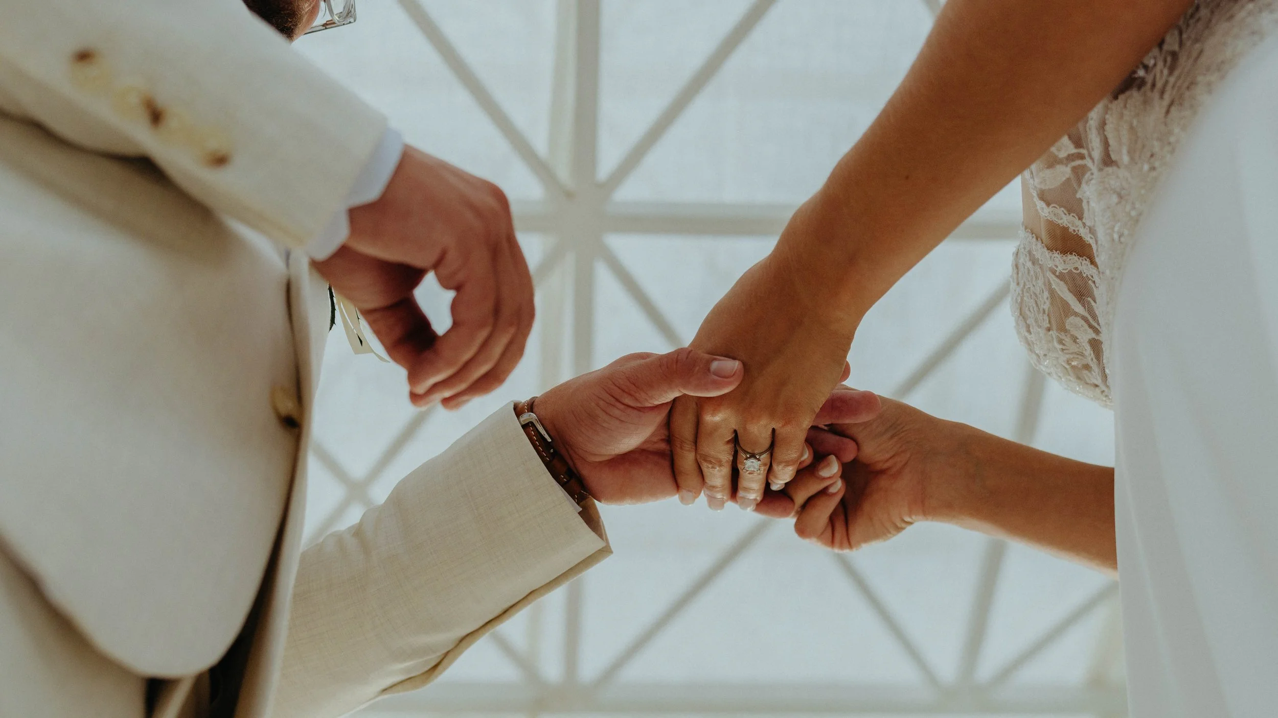 Close-up of a couple holding hands during a wedding ceremony, with the bride wearing a lace dress and the groom in a light-colored suit, under a white canopy.