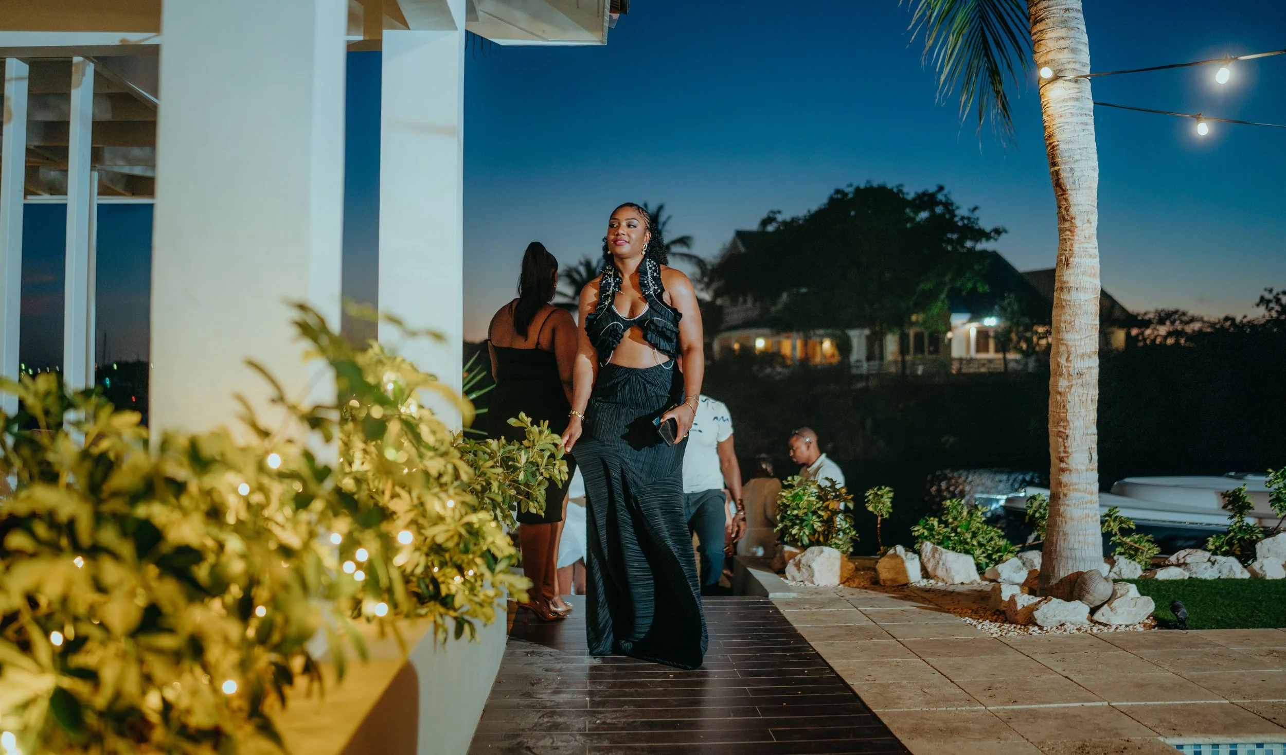 A woman in a black dress and matching top with earrings and bracelets stands outdoors during the evening, surrounded by other people and tropical plants with string lights and palm trees, under a darkening sky.