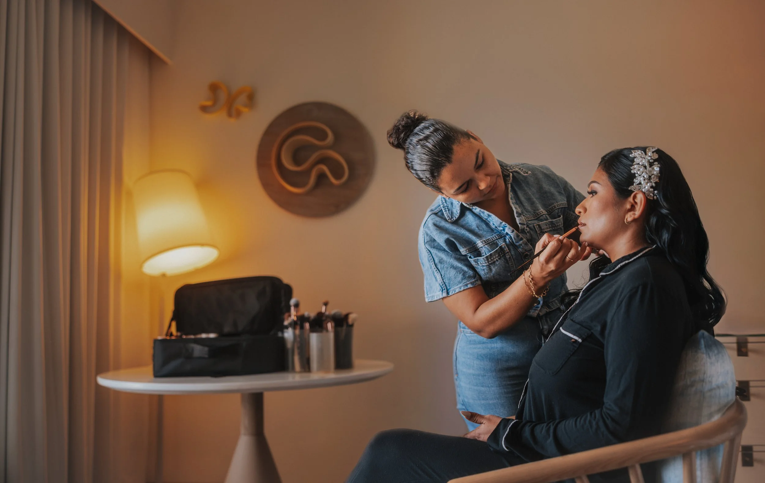 Makeup artist applying makeup to a woman with an ornate hairpiece, seated in a chair in a warmly lit room with a decorative wall art and a lamp.