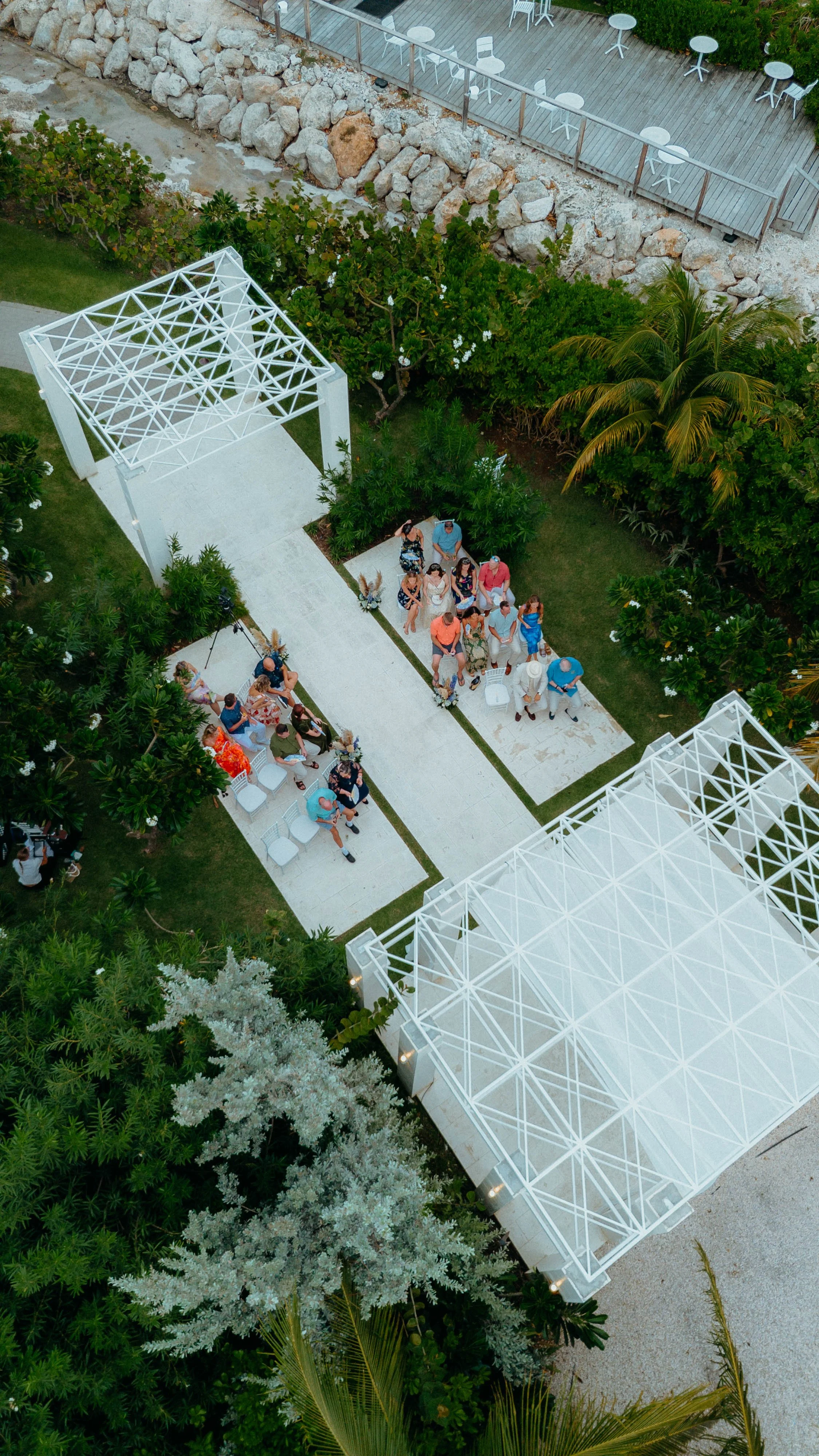 An aerial view of an outdoor event with people seated in chairs or standing on a pathway, lush green plants, trees, and a white canopy structure with a geometric pattern.
