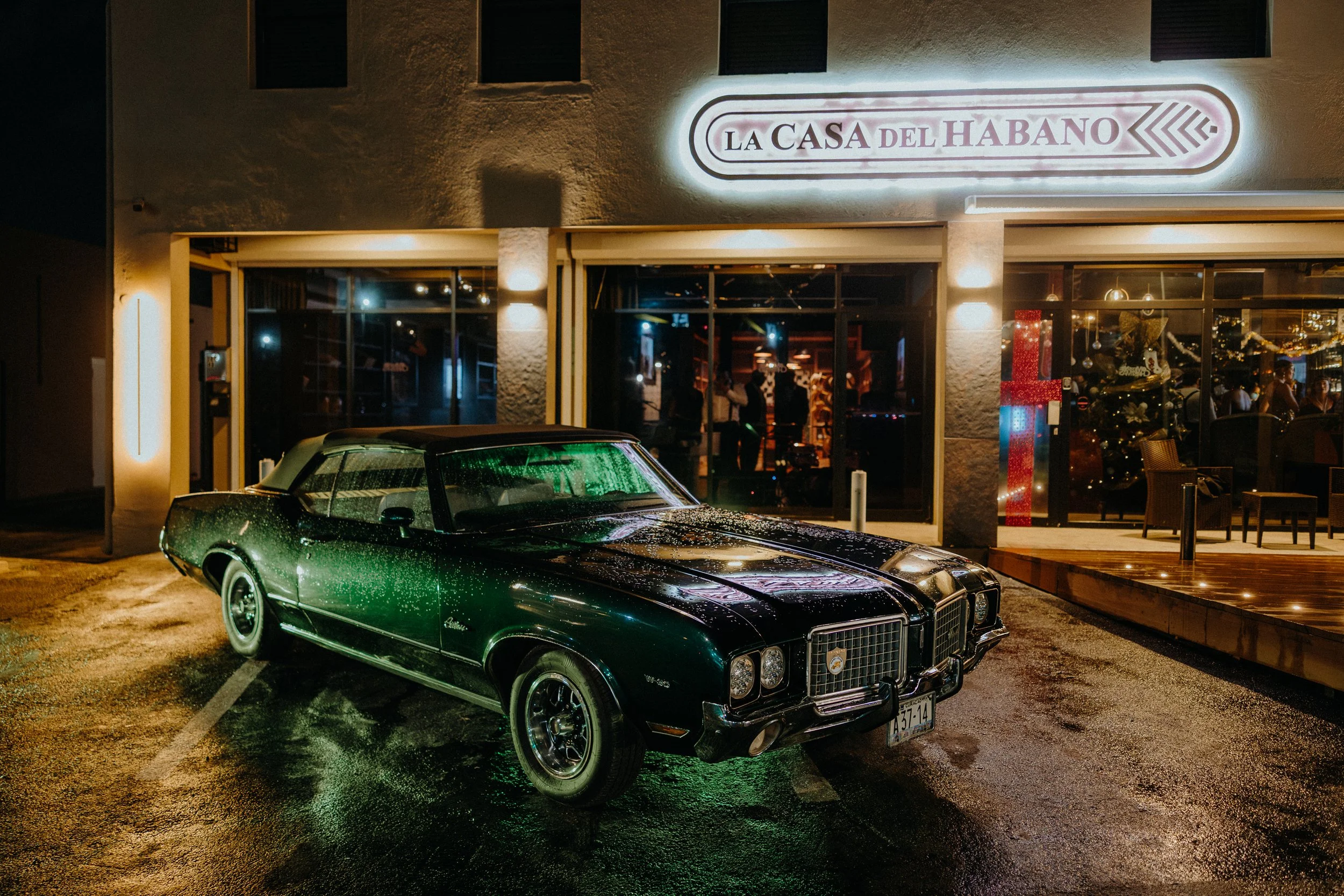 Nighttime scene outside a building with a red and white sign reading 'La Casa del Habano.' A vintage green car with water droplets on its surface is parked in front. Inside, festive holiday decorations and people are visible through the large windows