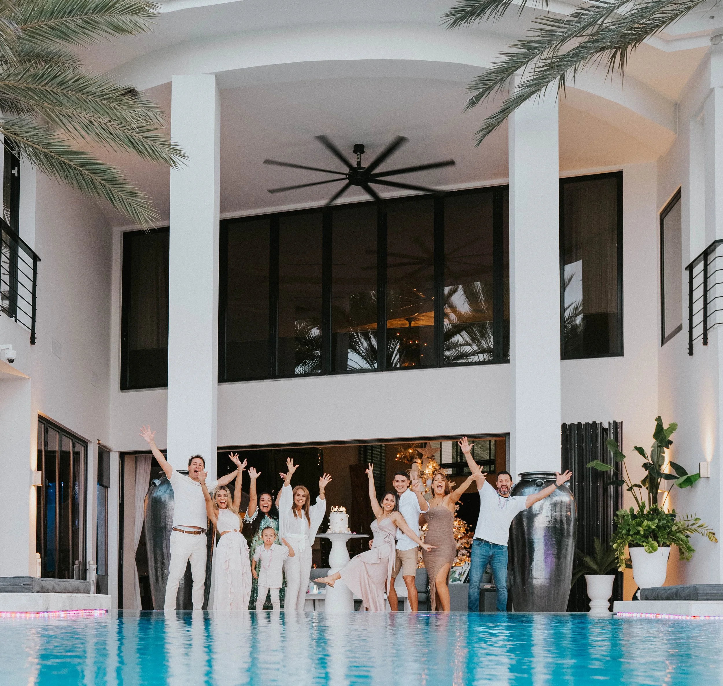 Group of people celebrating a birthday party by a swimming pool in a modern house with large windows and white walls. They are smiling, raising their hands, and some are jumping. There is a birthday cake on a small table.