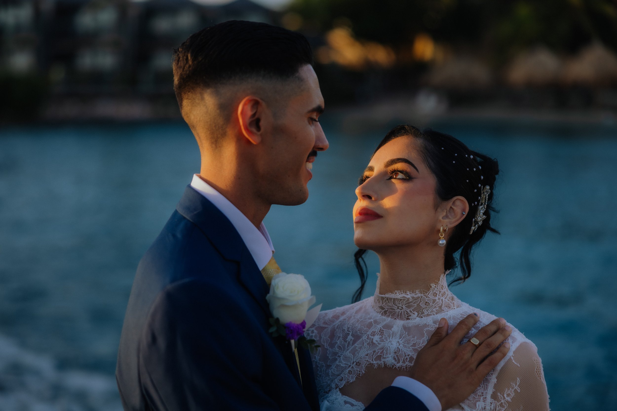 A newlywed couple dancing outdoors at sunset near a body of water, with the groom smiling and the bride looking up at him.