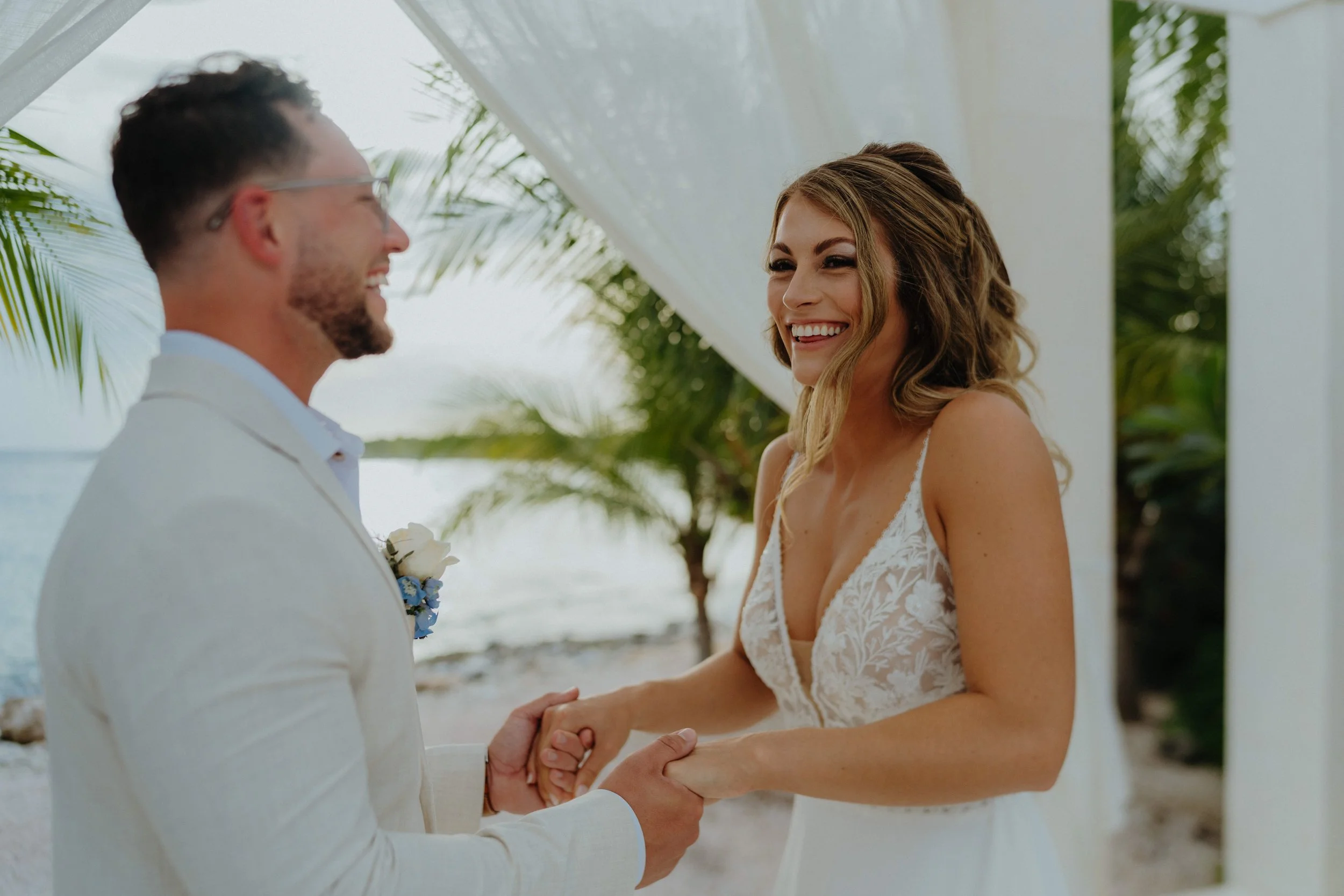 A bride and groom holding hands and smiling during their wedding ceremony outdoors, with tropical plants and a white canopy in the background.