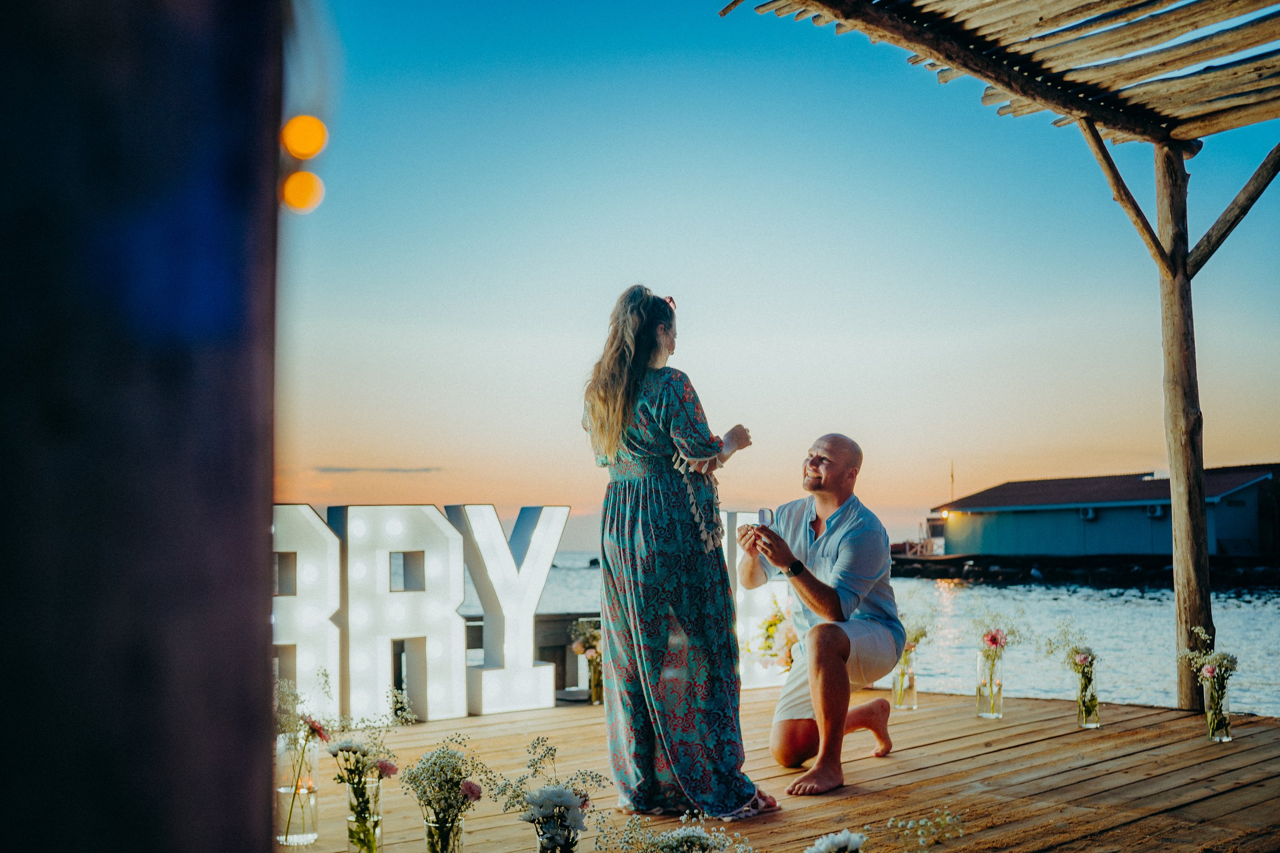 A woman in a long colorful dress is proposing to a man on a beach at sunset. The man is kneeling and holding out a ring, smiling. In the background, there are decorative flowers, a large illuminated sign that reads 'LOVE,' and a rustic wooden structure. The sky is clear and the ocean is visible.