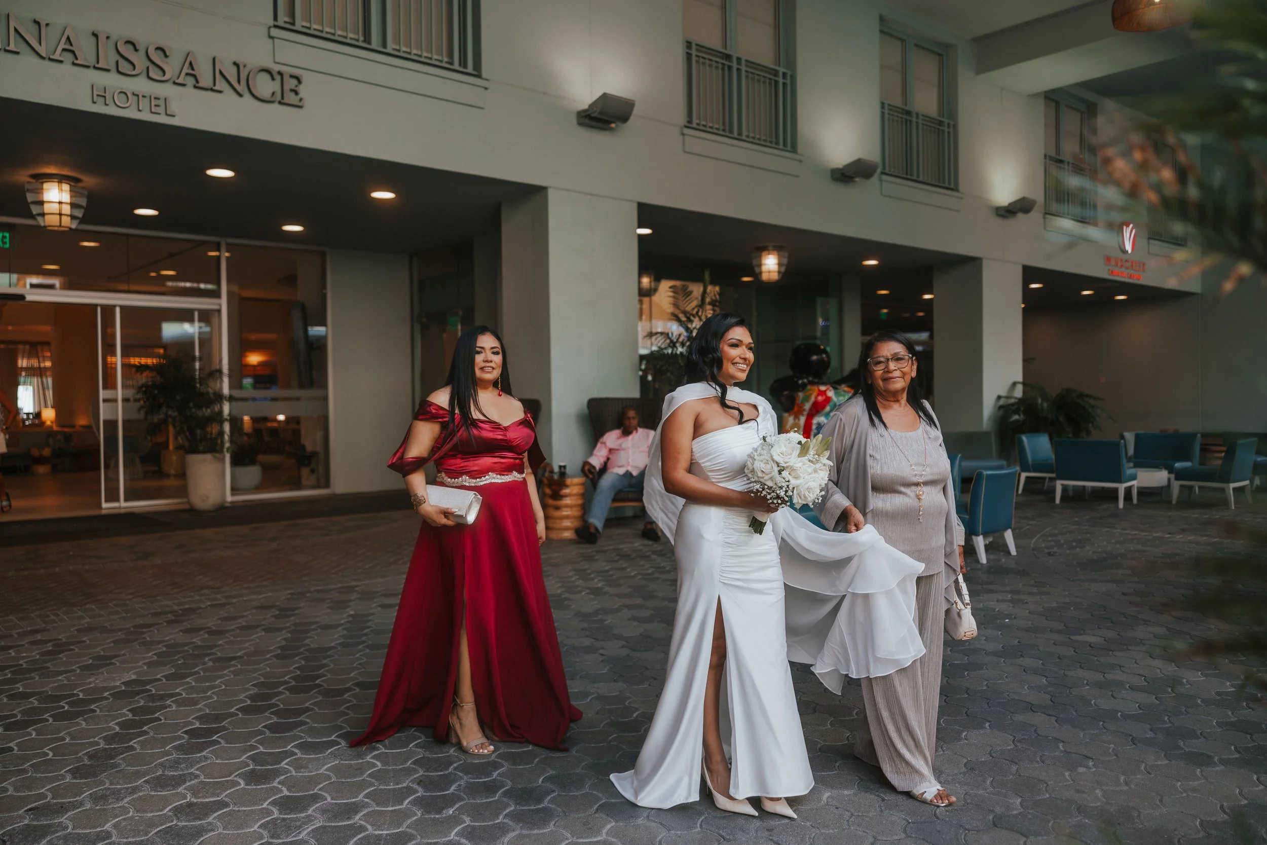 A bride in a white wedding dress holding a bouquet walking with two women outside a hotel, with a hotel entrance and a sign that reads 'NAISSANCE HOTEL' in the background.