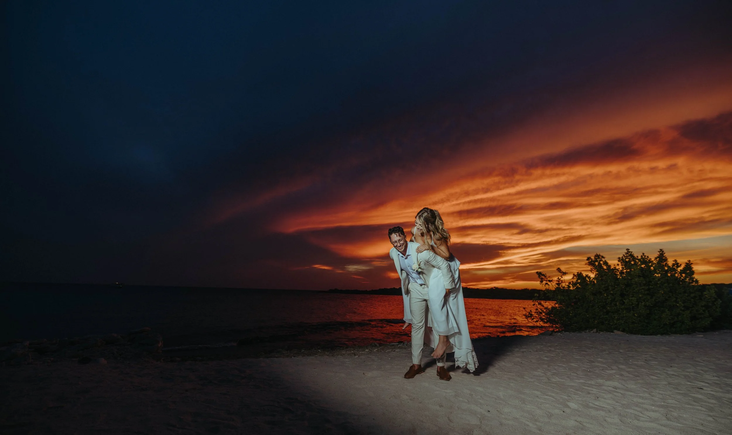 A couple in wedding attire at sunset by the water, with the groom carrying the bride on his back, smiling and enjoying the moment.
