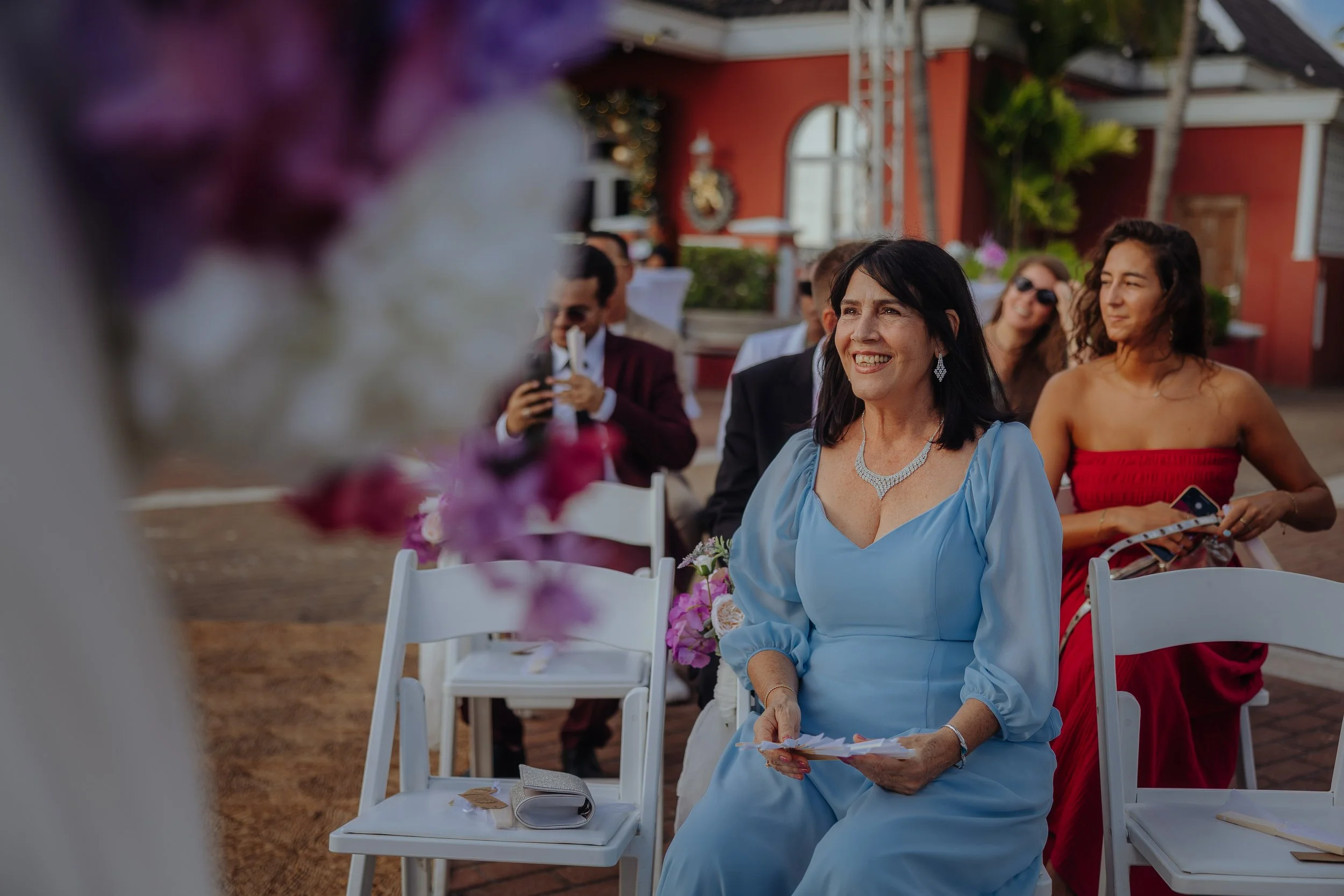 A woman in a light blue dress sitting at an outdoor event, smiling, surrounded by other guests, with a woman in a red dress and sunglasses sitting behind her.