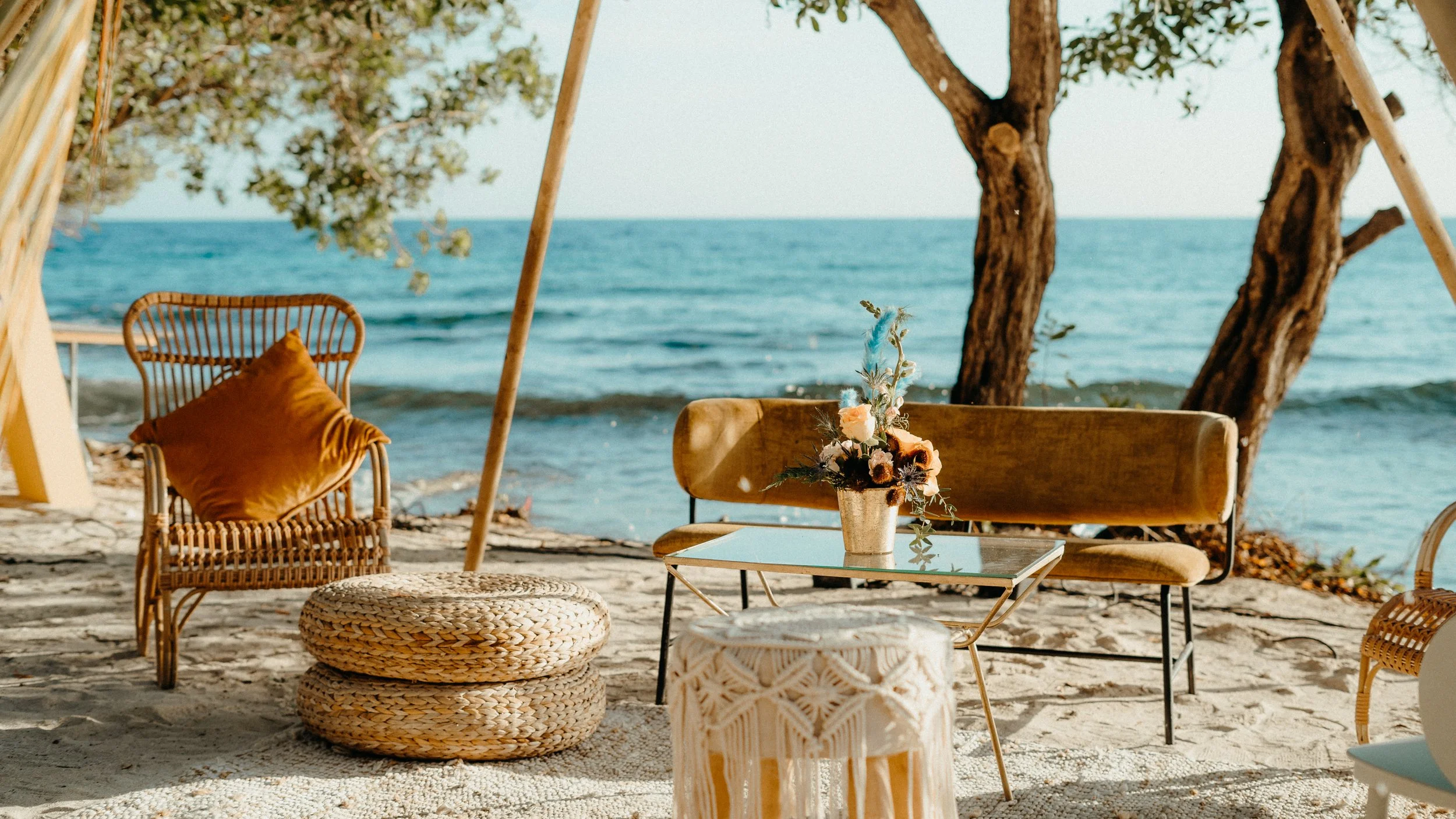 Beachside seating area with rattan chairs, a coffee table with a flower vase, and woven ottomans, overlooking the ocean with trees in the background.