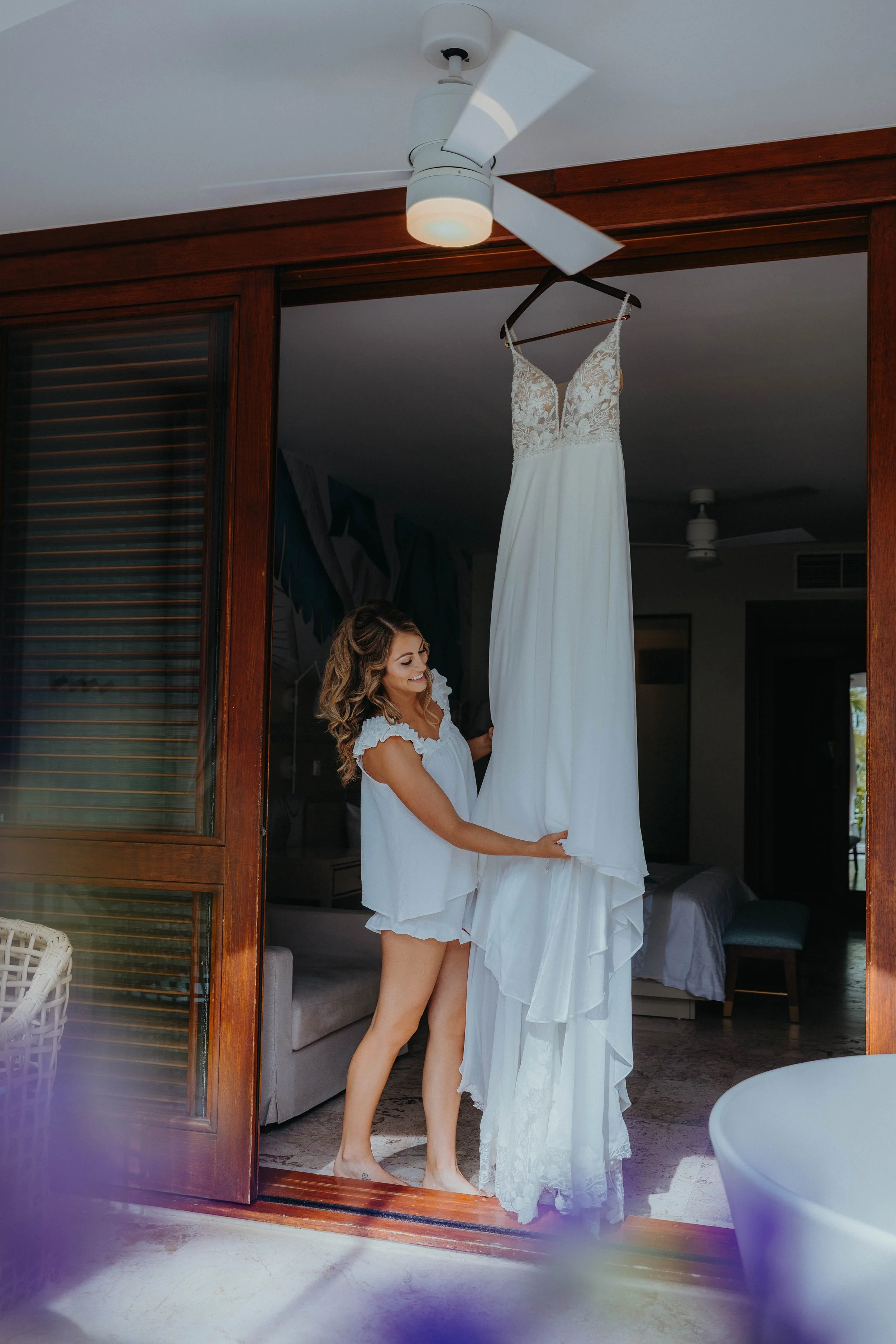 A woman in a white nightgown hanging a white wedding dress on a hanger inside a room with wooden sliding door frame, overlooking a bed and living area.