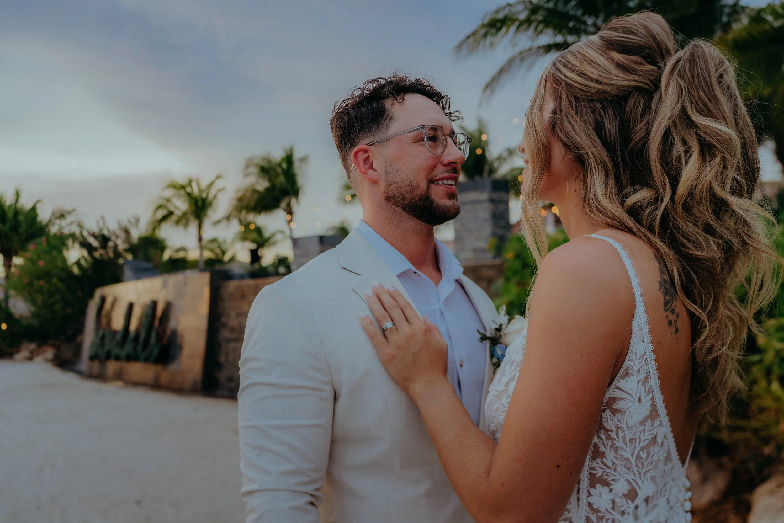 A groom and bride face each other closely at sunset outdoors, with palm trees and string lights in the background.